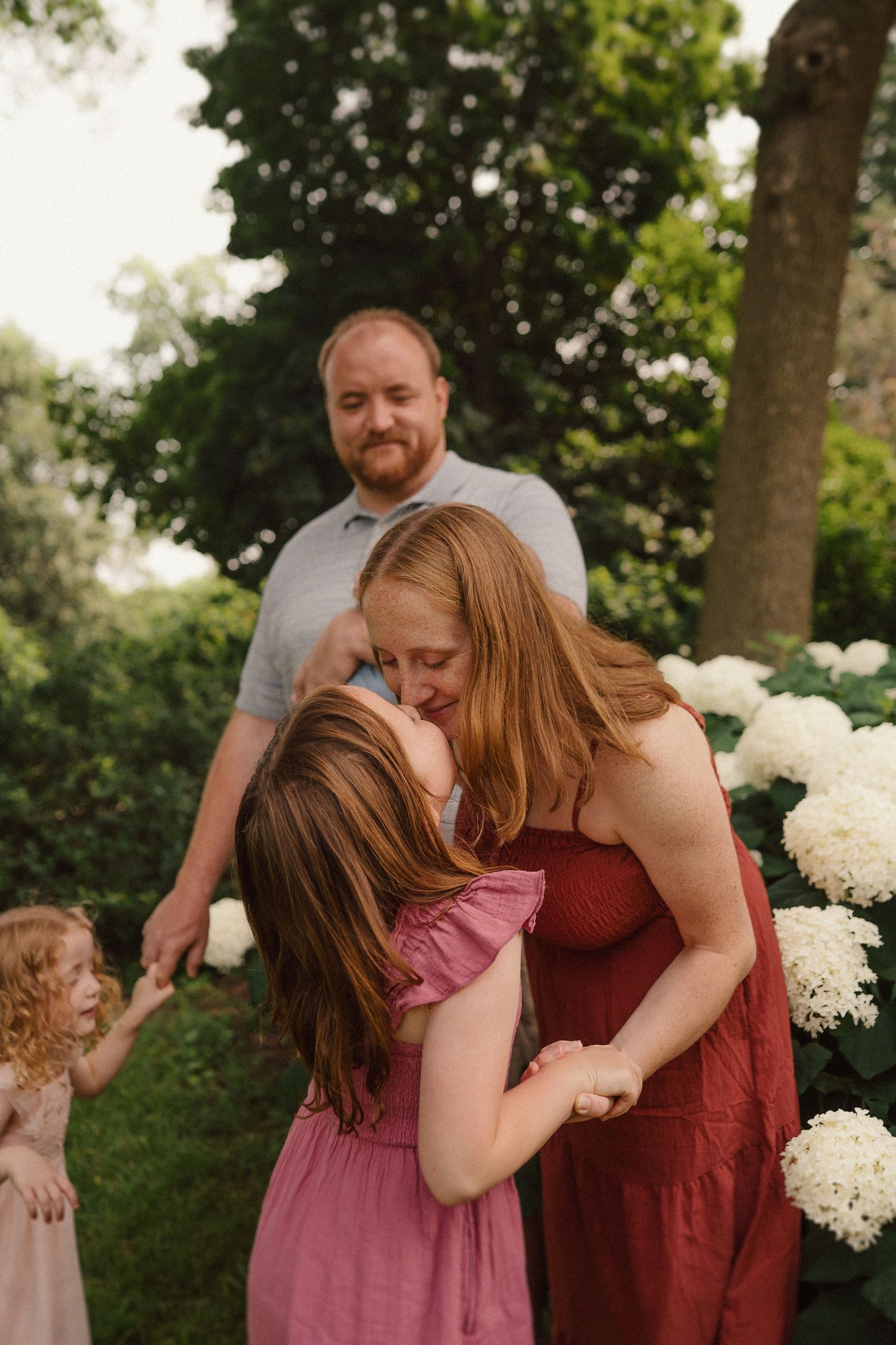 A tender family moment captured in a garden setting with blooming white hydrangeas in the background.