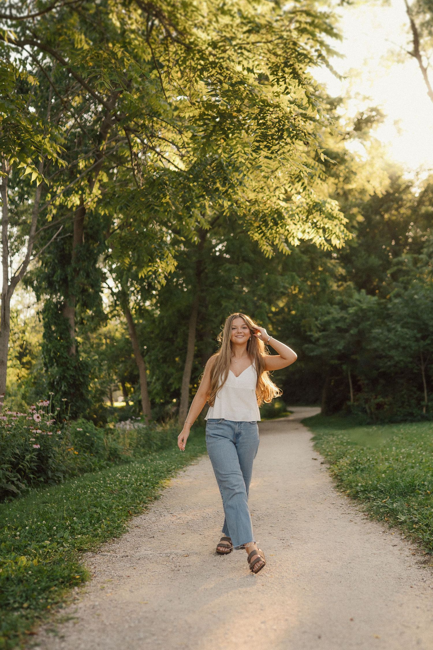 A woman in casual attire strolls down a tree-lined path during sunset.