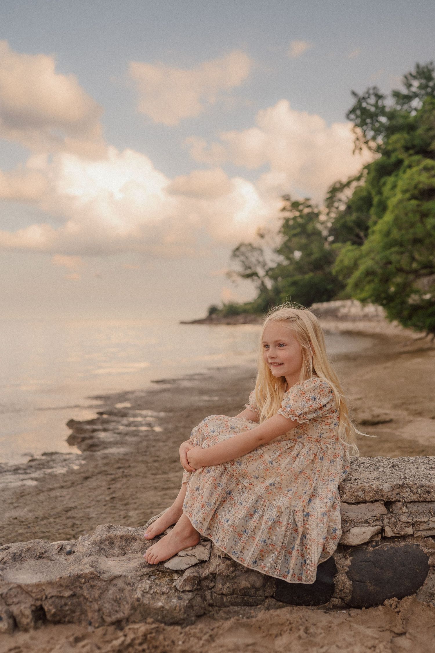 Child in floral dress sits peacefully on rocky beach shore during golden sunset hour with trees in background.
