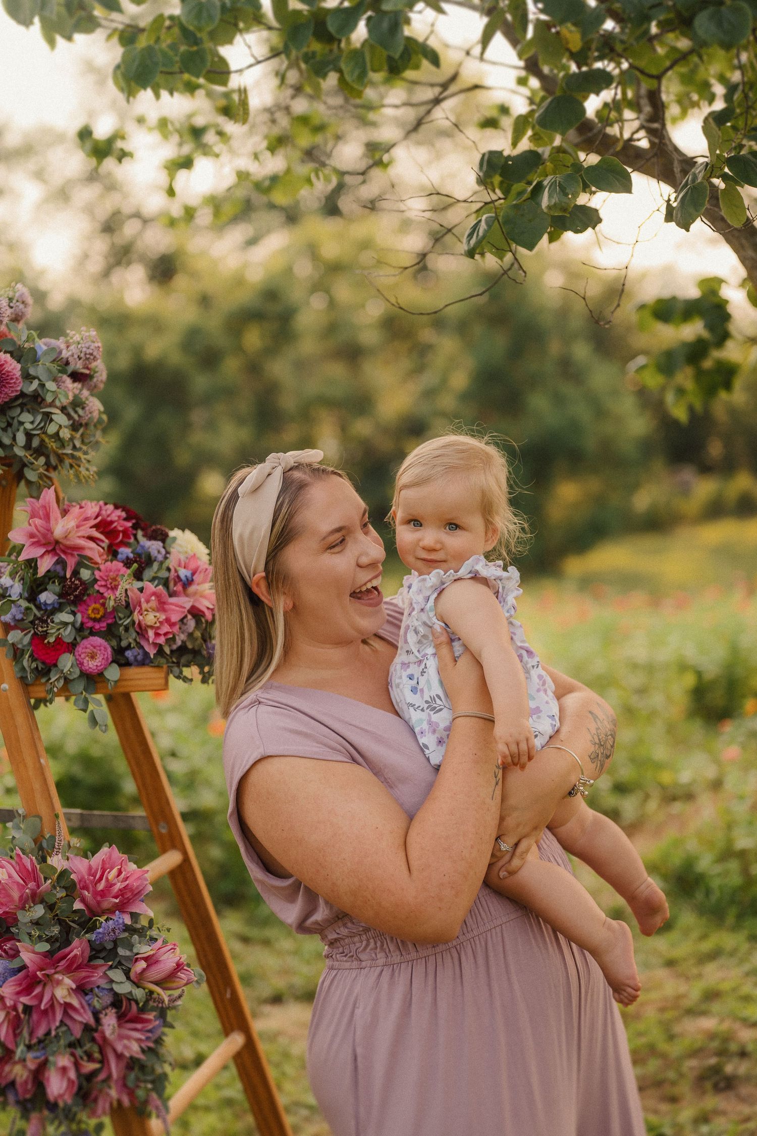 A woman in a mauve dress shares a joyful moment with a small child against a backdrop of floral decorations outdoors.