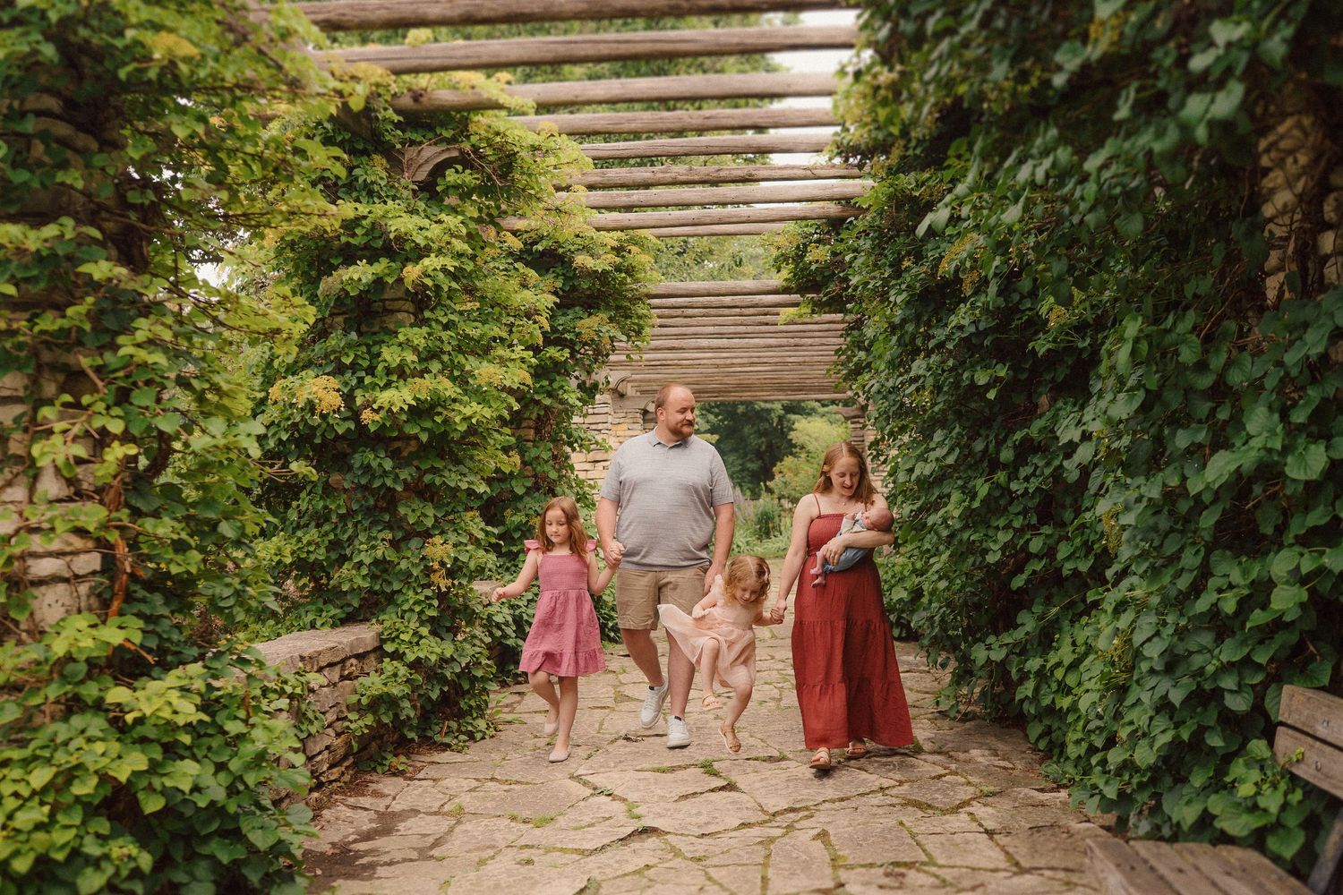 A family walks together along a beautiful garden path under a wooden pergola covered in lush greenery.