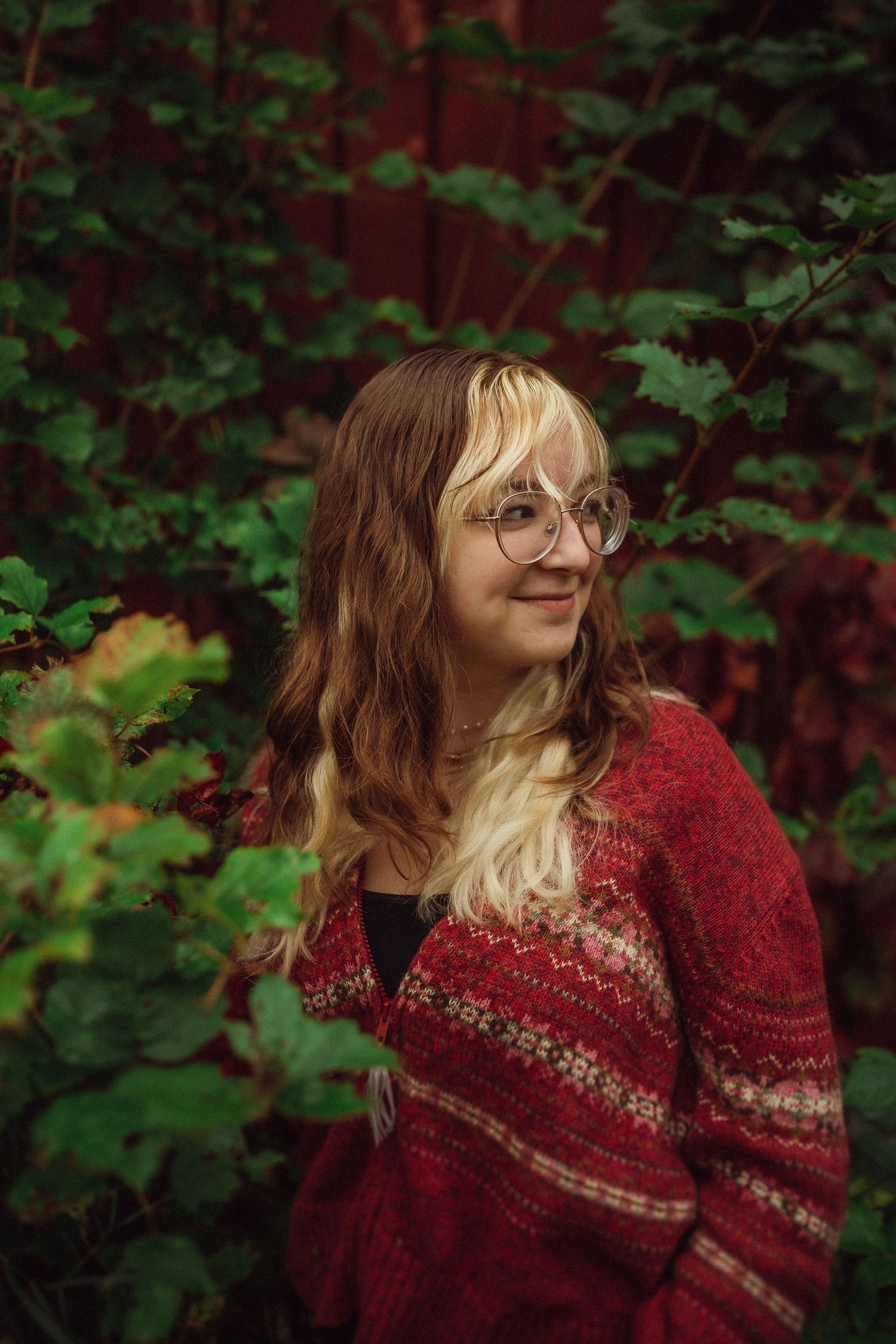 An autumn portrait featuring a red patterned sweater against lush green foliage and rustic red siding.