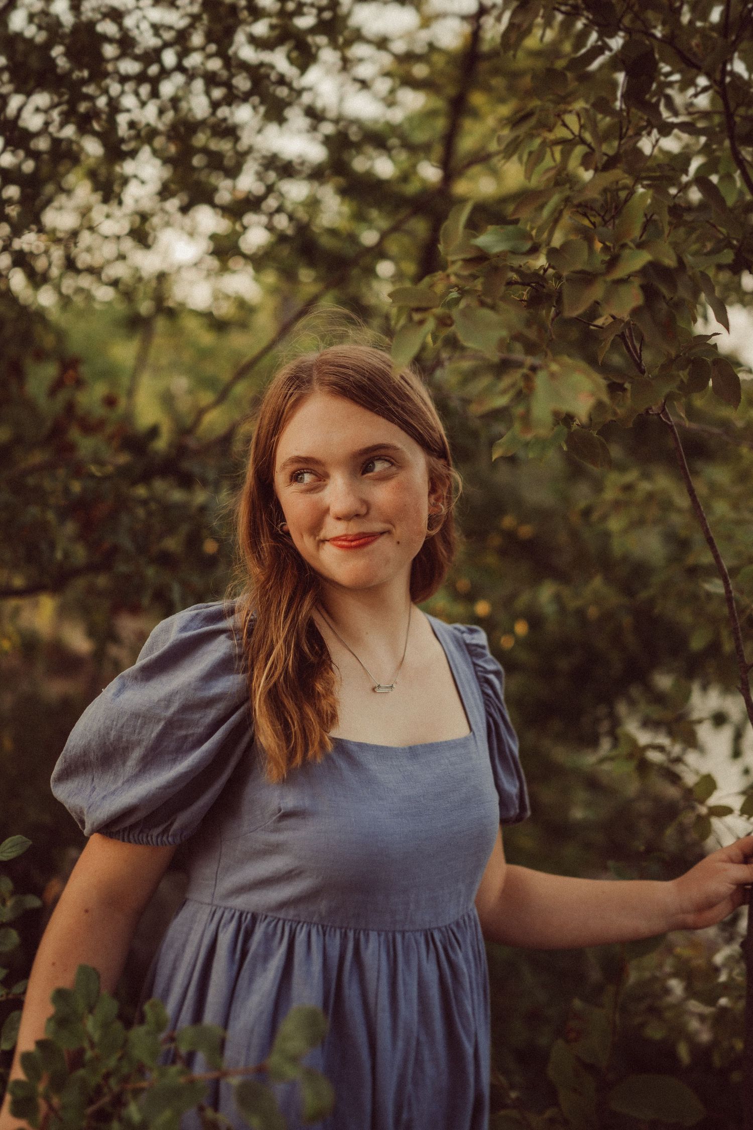 A portrait in dappled sunlight showing someone in a blue dress with puff sleeves among tree branches.