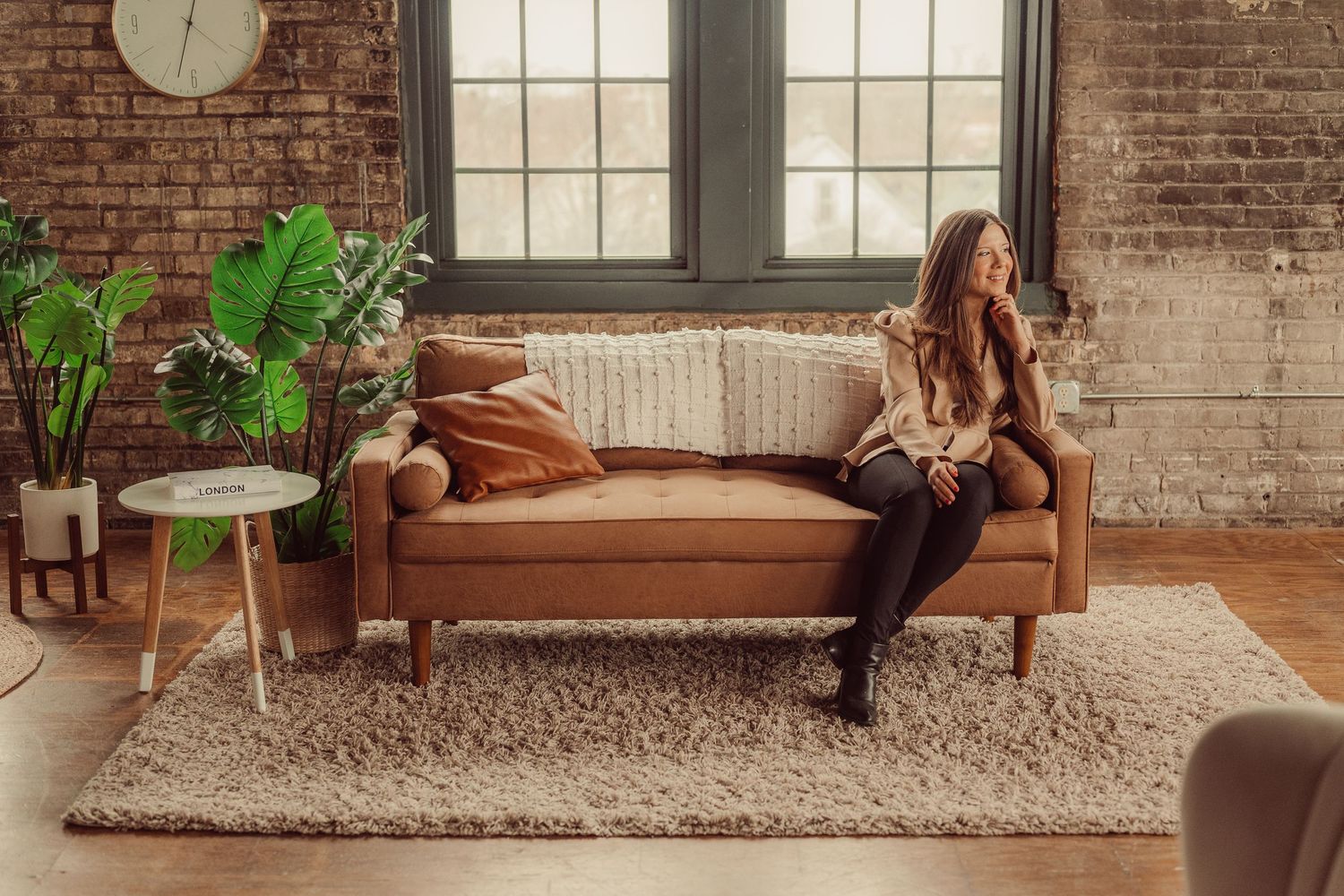 Contemporary living room featuring camel leather sofa against textured brick wall with large windows.