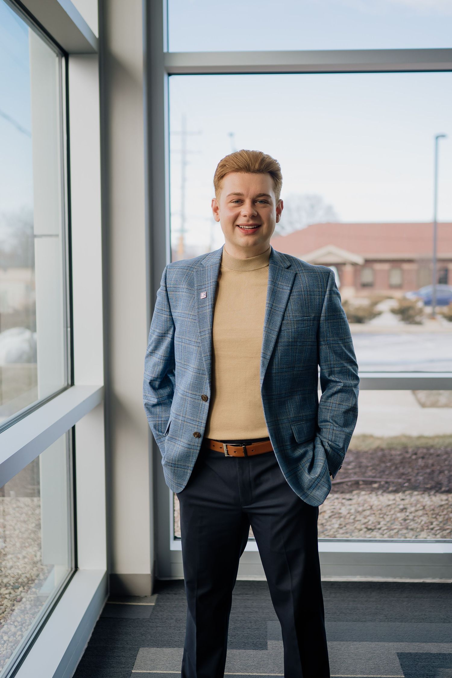 Professional standing portrait by window wearing blue blazer and tan turtleneck with natural lighting.