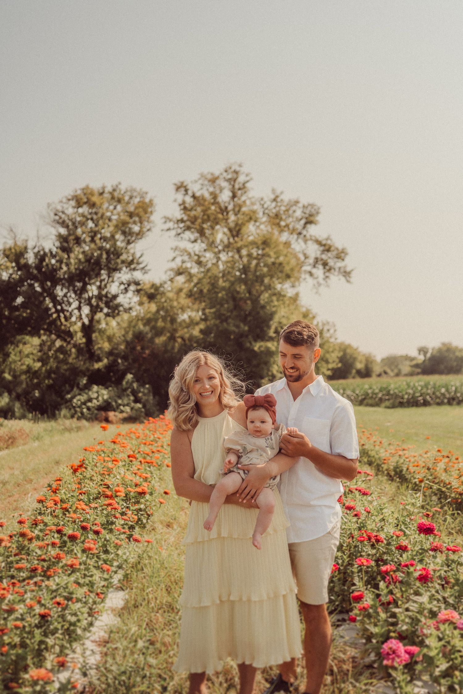 Couple stands with infant in a field of vibrant red flowers during golden hour sunset.