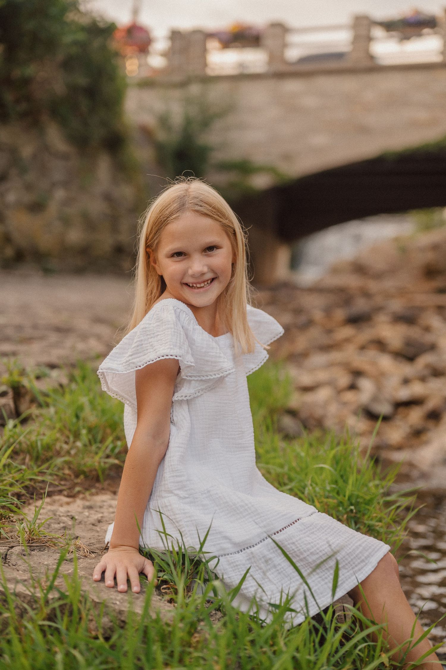 A child in a white dress plays in grass near rocks outdoors.