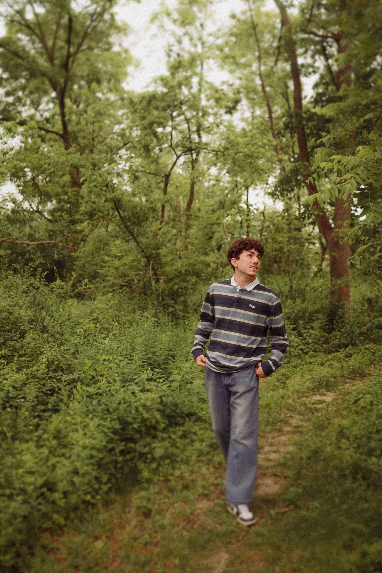 A person in a striped sweater and jeans walks along a wooded trail surrounded by lush green foliage on a summer day.