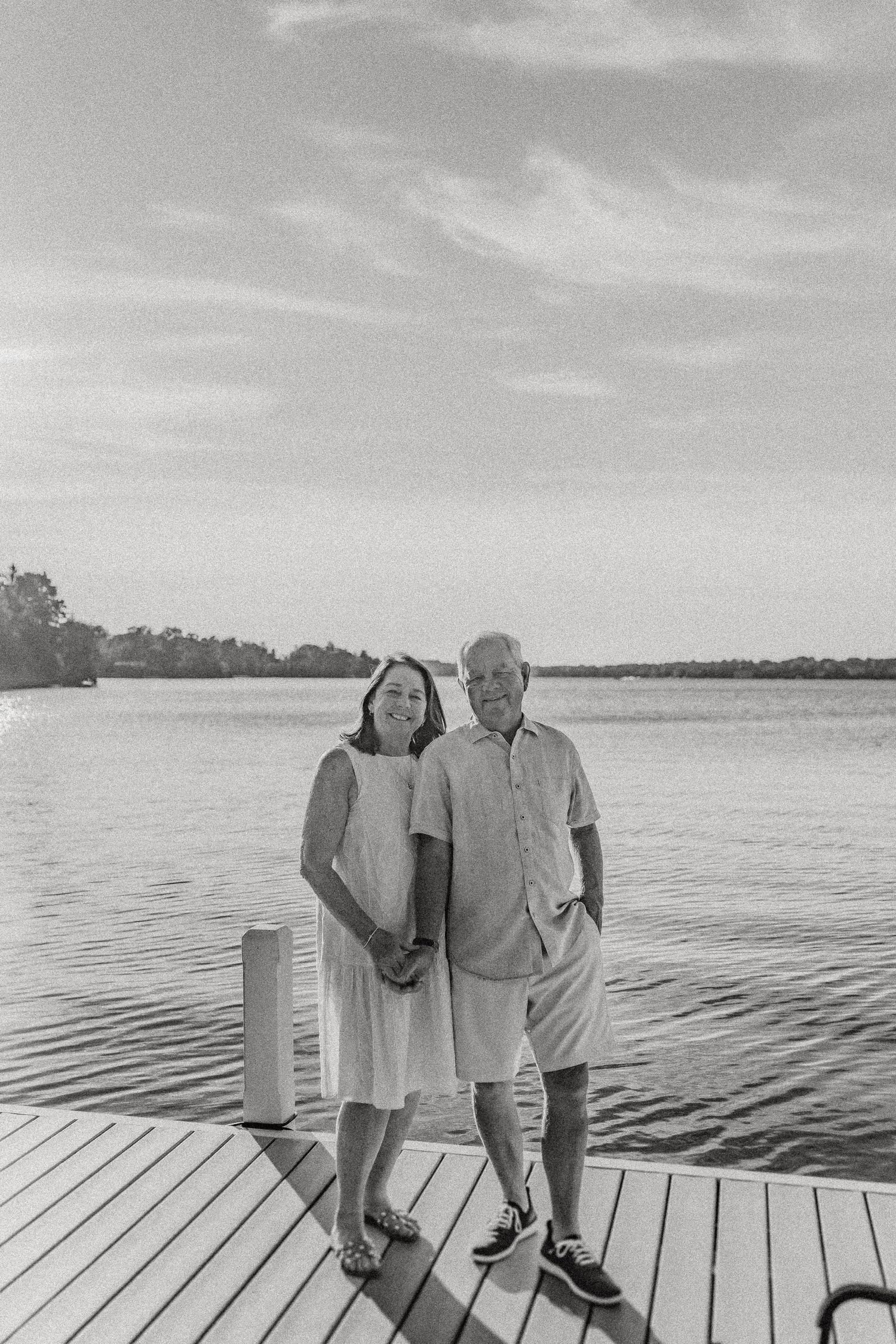 A couple stands together on a wooden dock overlooking a peaceful lake at sunset.