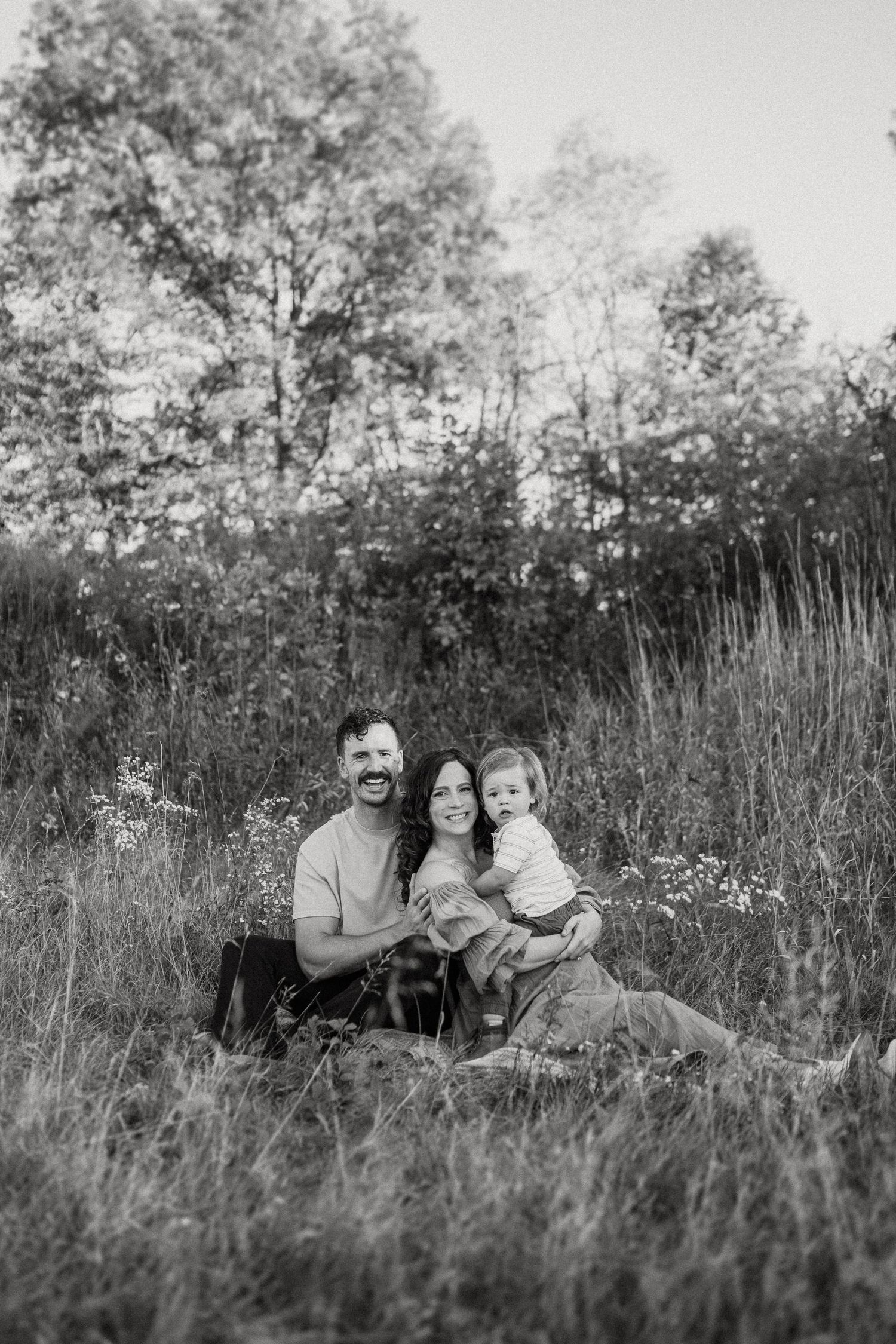 Black and white portrait of a family sitting together in a meadow with trees in the background.