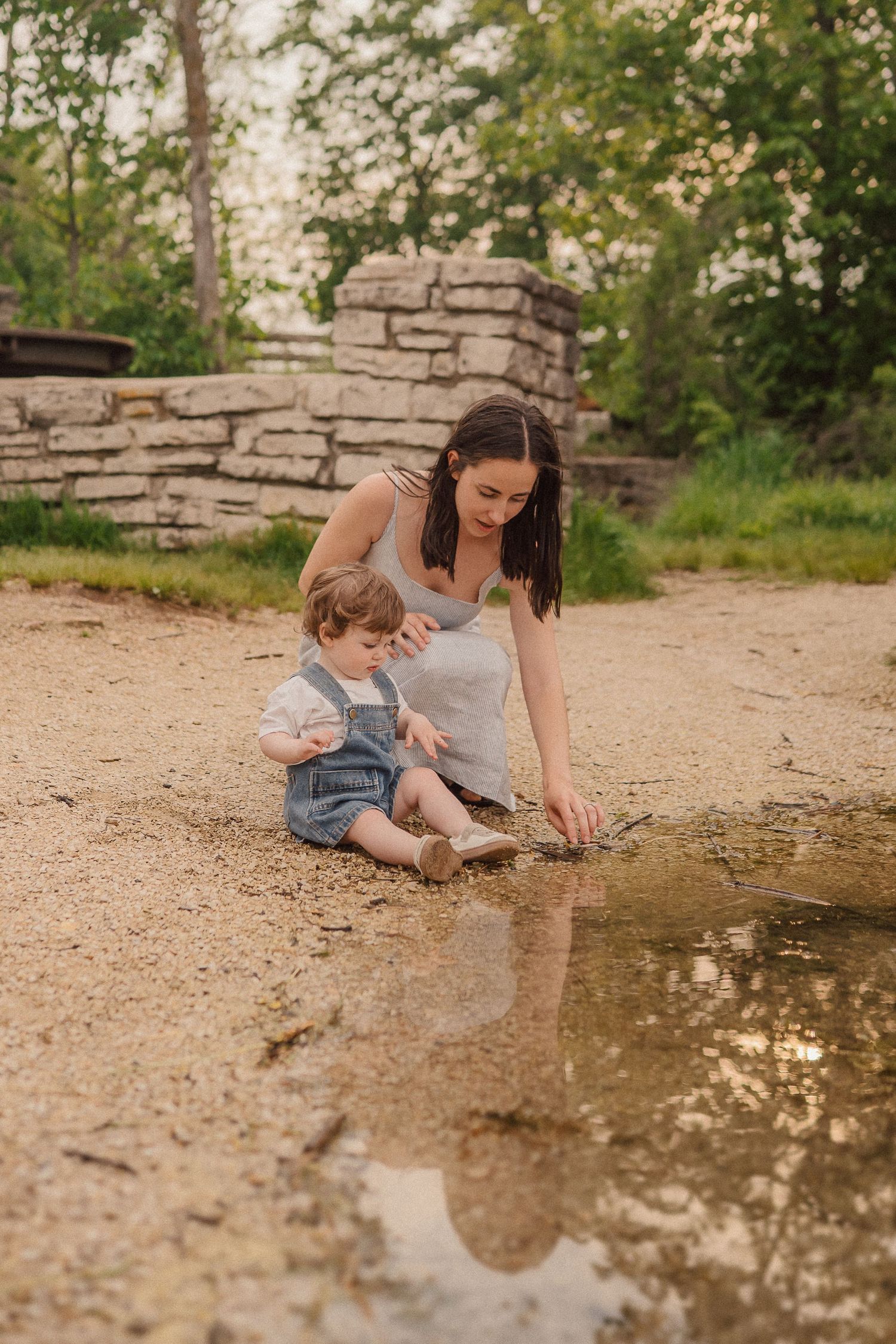 A tender moment by a puddle on a dirt path with a stone wall and trees in the background.
