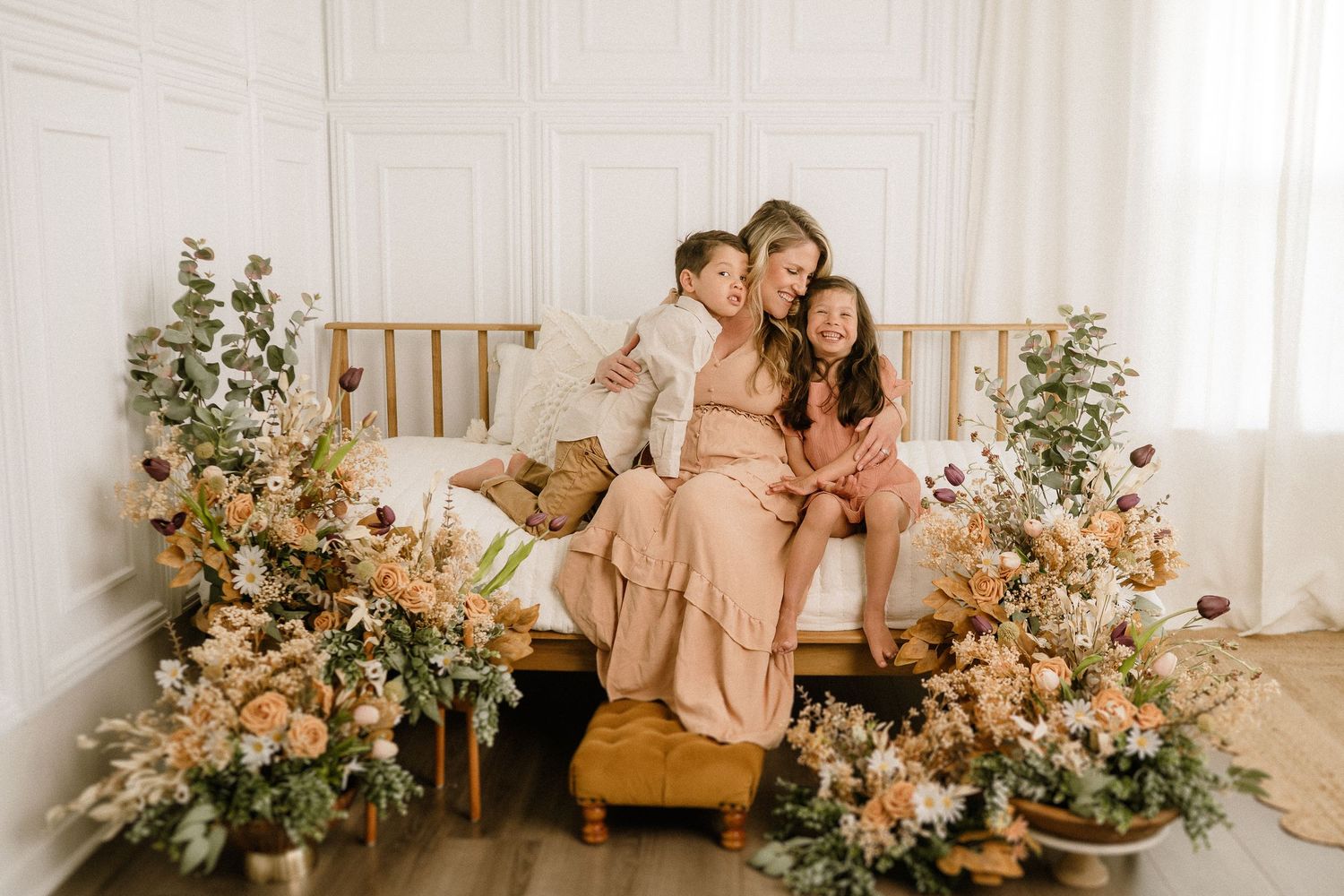 Two women in neutral-toned dresses sit on a gold bed frame surrounded by romantic floral arrangements in a bright room.