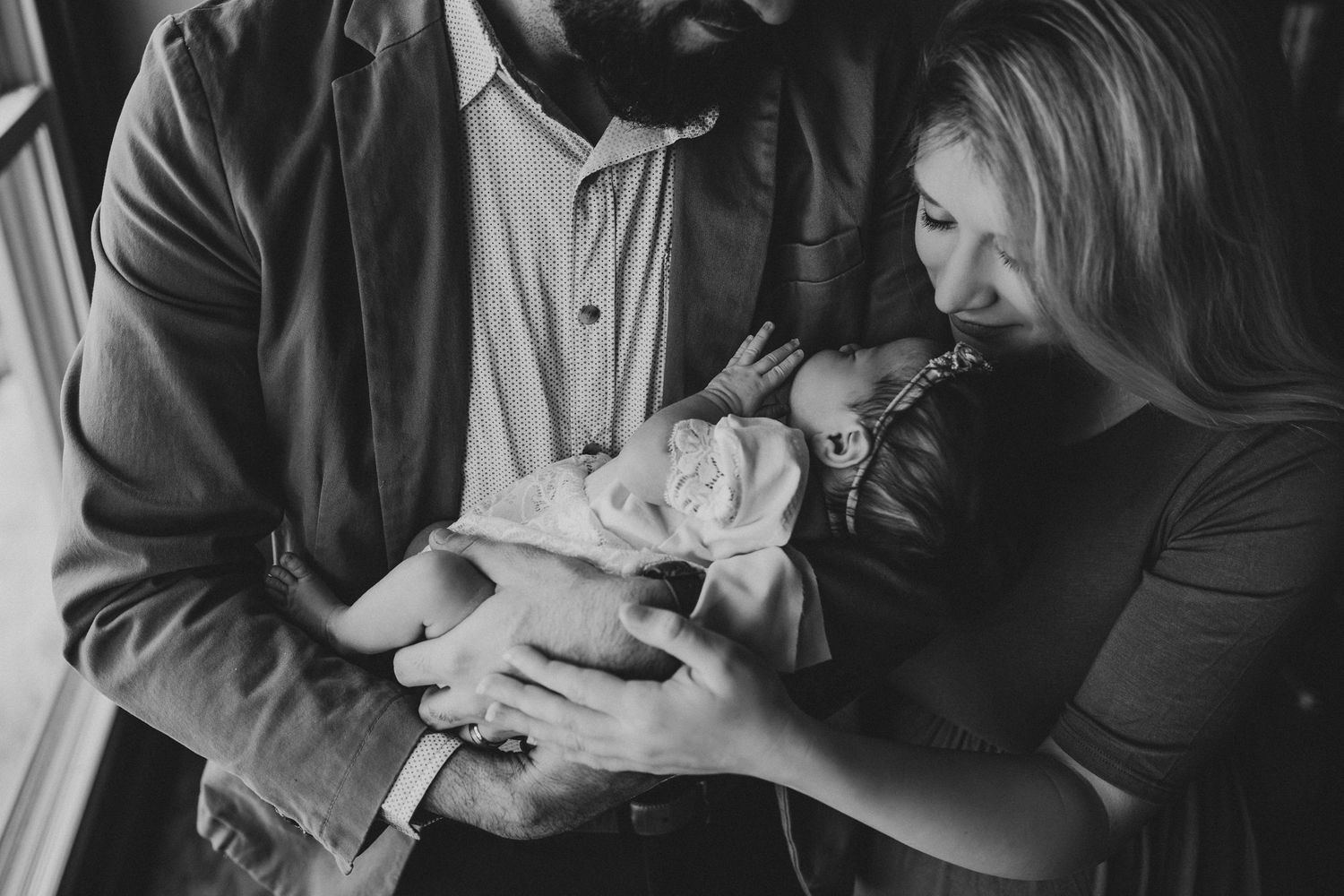 Black and white image of a couple sharing an intimate moment with their newborn by a window.