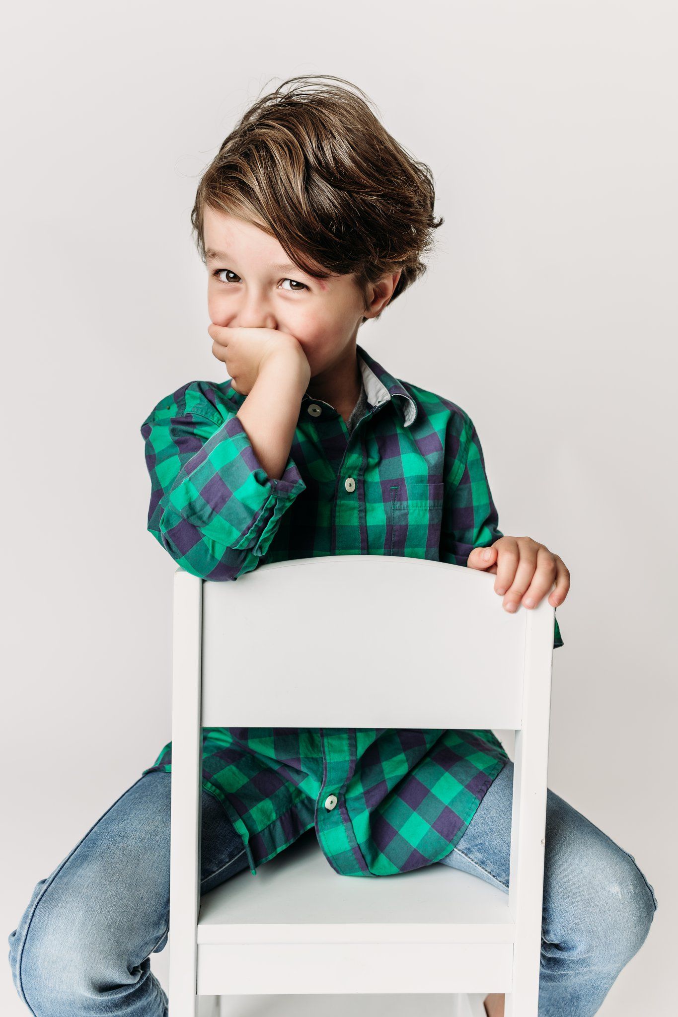 Child in green plaid shirt sits casually on white chair against neutral background.