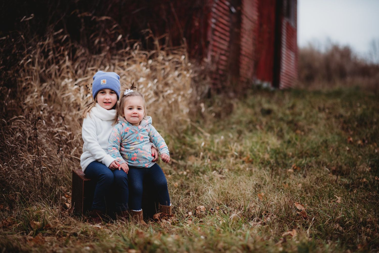 Two small children sit together on a wooden bench against a rustic red barn backdrop.