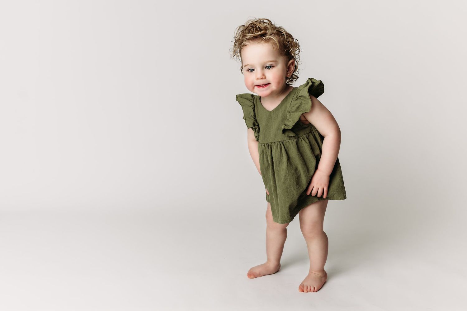 Child in olive green ruffle dress stands against white background.