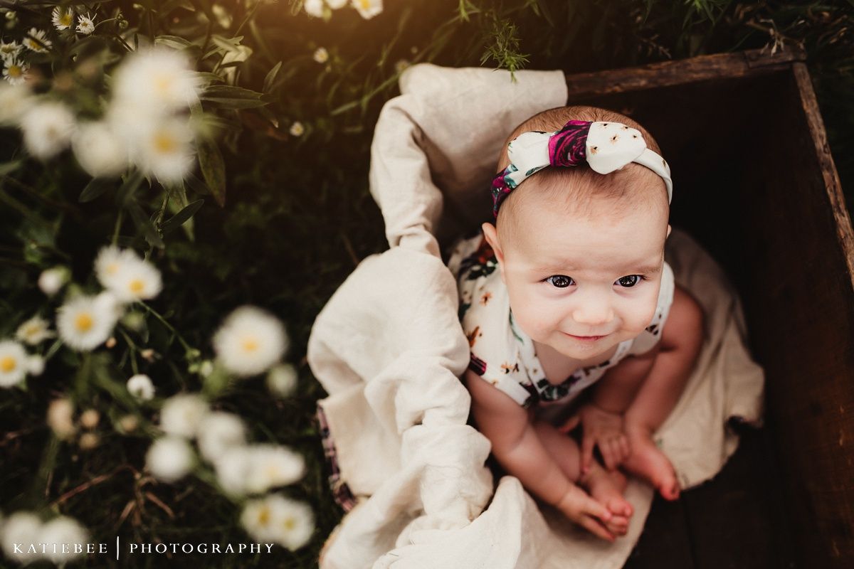 Baby wearing white bow sits in wooden box surrounded by delicate white flowers.