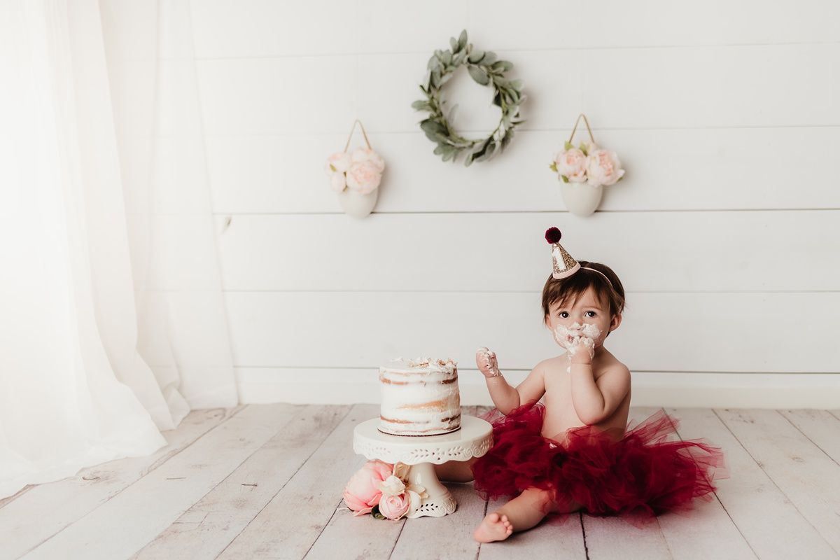 Baby in red tutu sits by birthday cake with wreath and decorations on white shiplap wall.