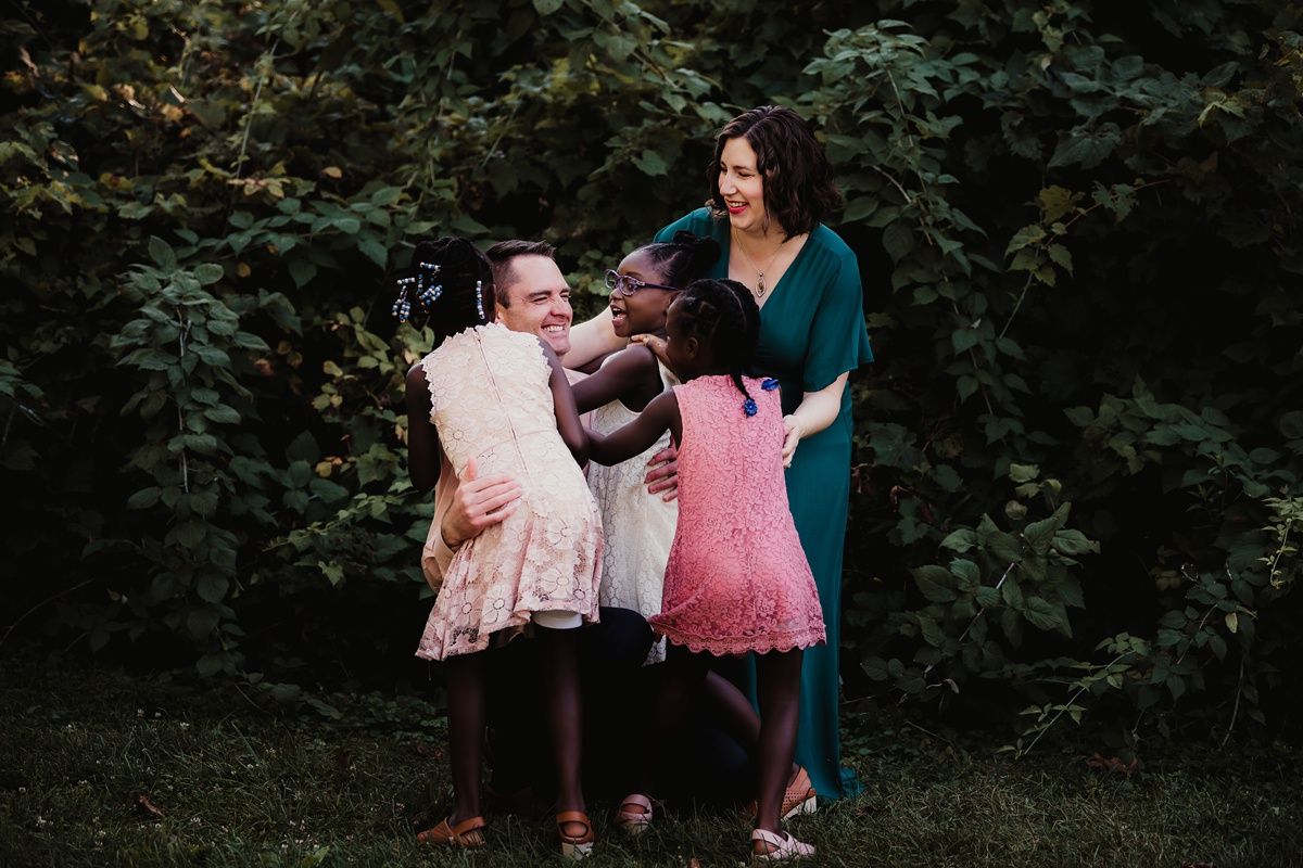 Family portrait in garden setting with teal and pink dresses among greenery.