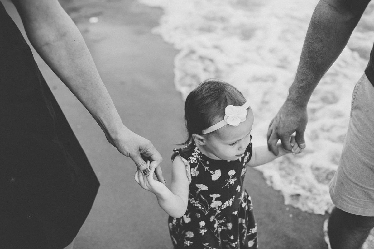 A black and white beach scene with tiny hands being held as waves roll in.