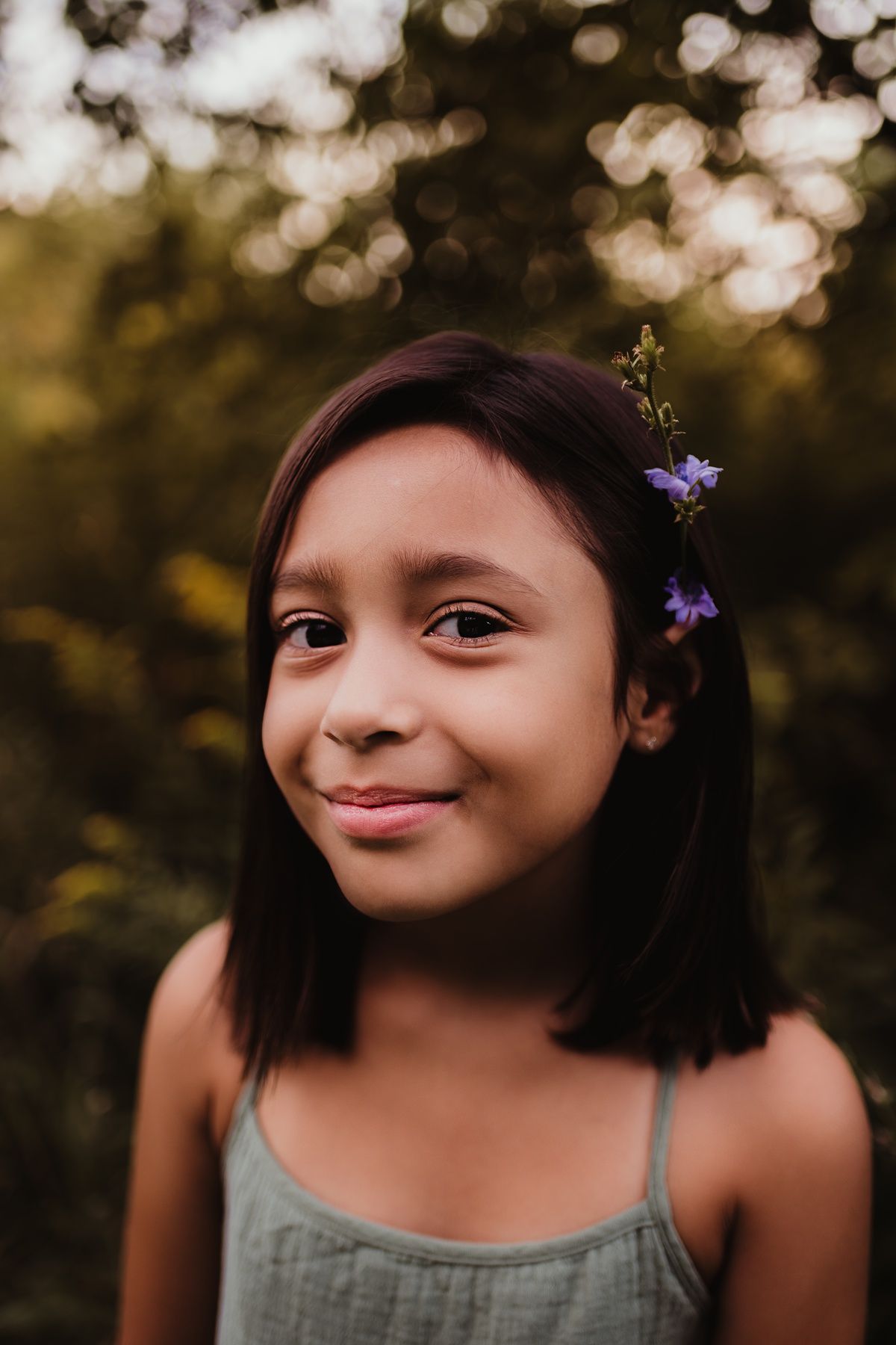 Portrait showing a gentle smile with purple flowers tucked behind the ear in golden light.