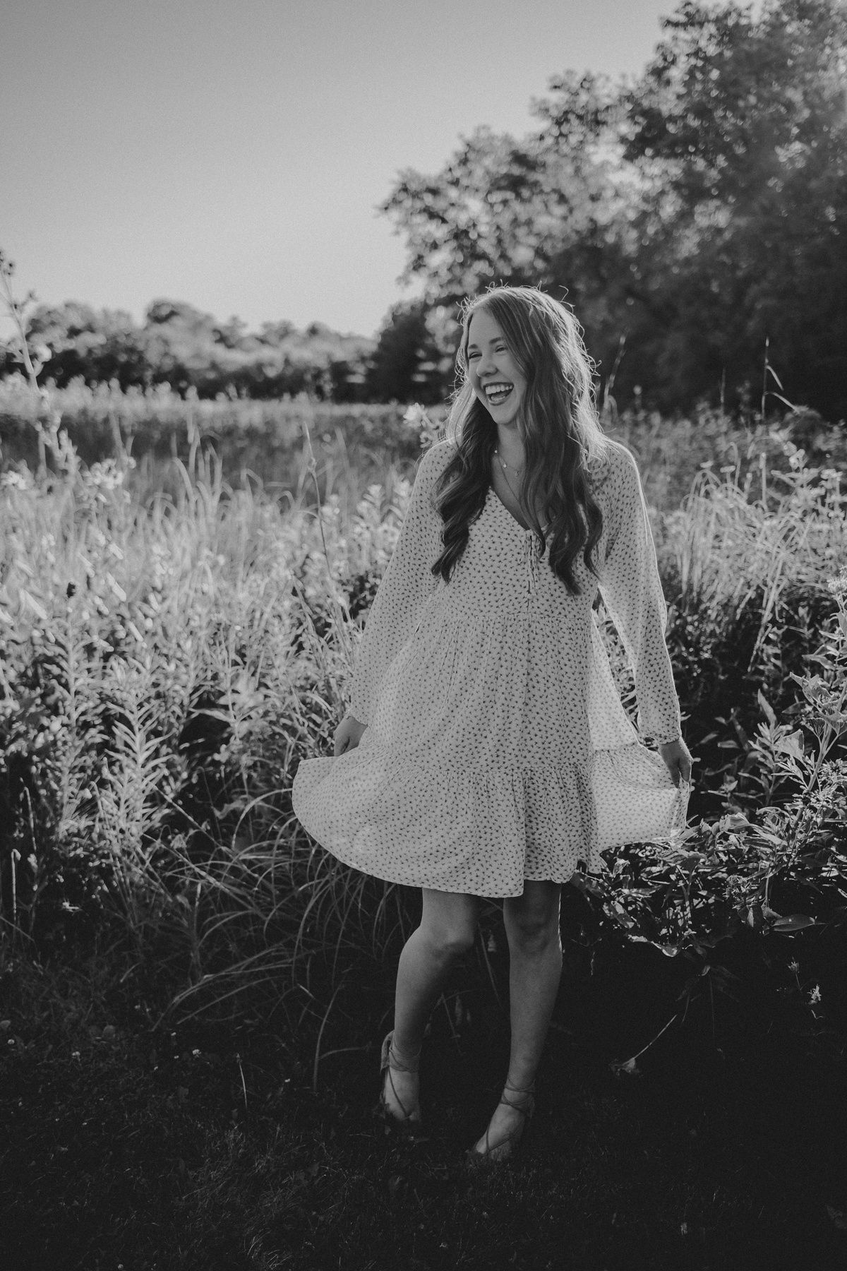 Black and white photo of someone laughing in a dotted dress amidst a field of wildflowers at sunset.