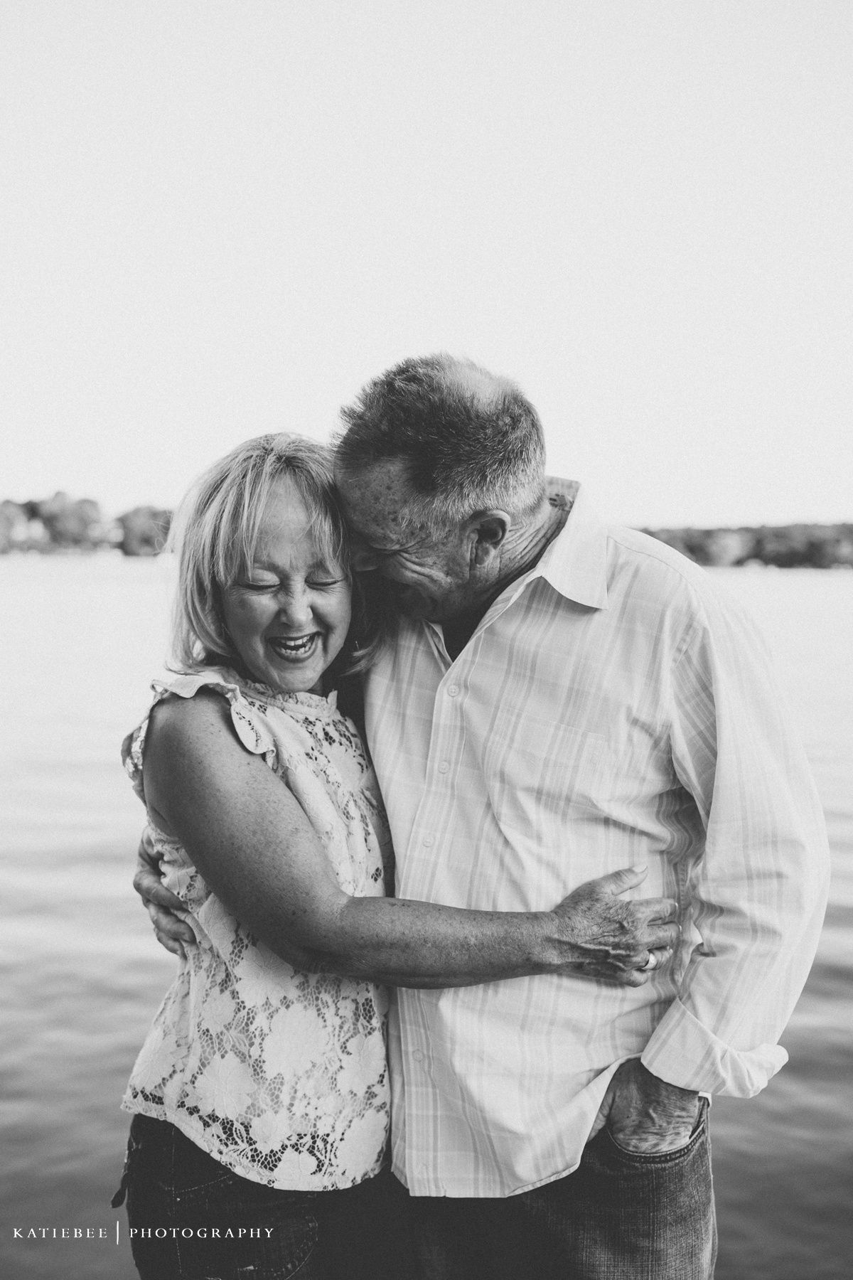 Black and white portrait of a couple sharing an intimate laughing moment by the water.