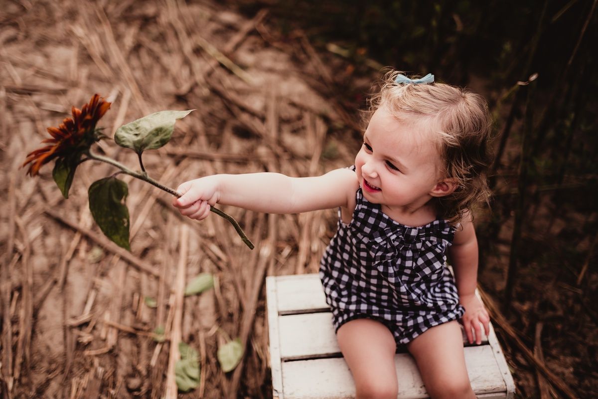 Child in polka dot dress sitting on vintage crate reaching for sunflower.