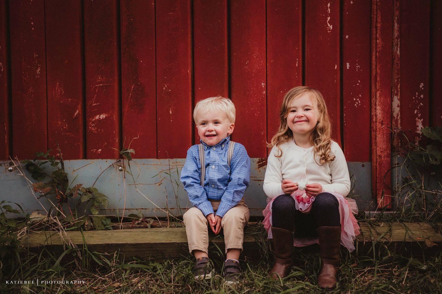 Two children sitting together on grass in front of a weathered red barn wall.