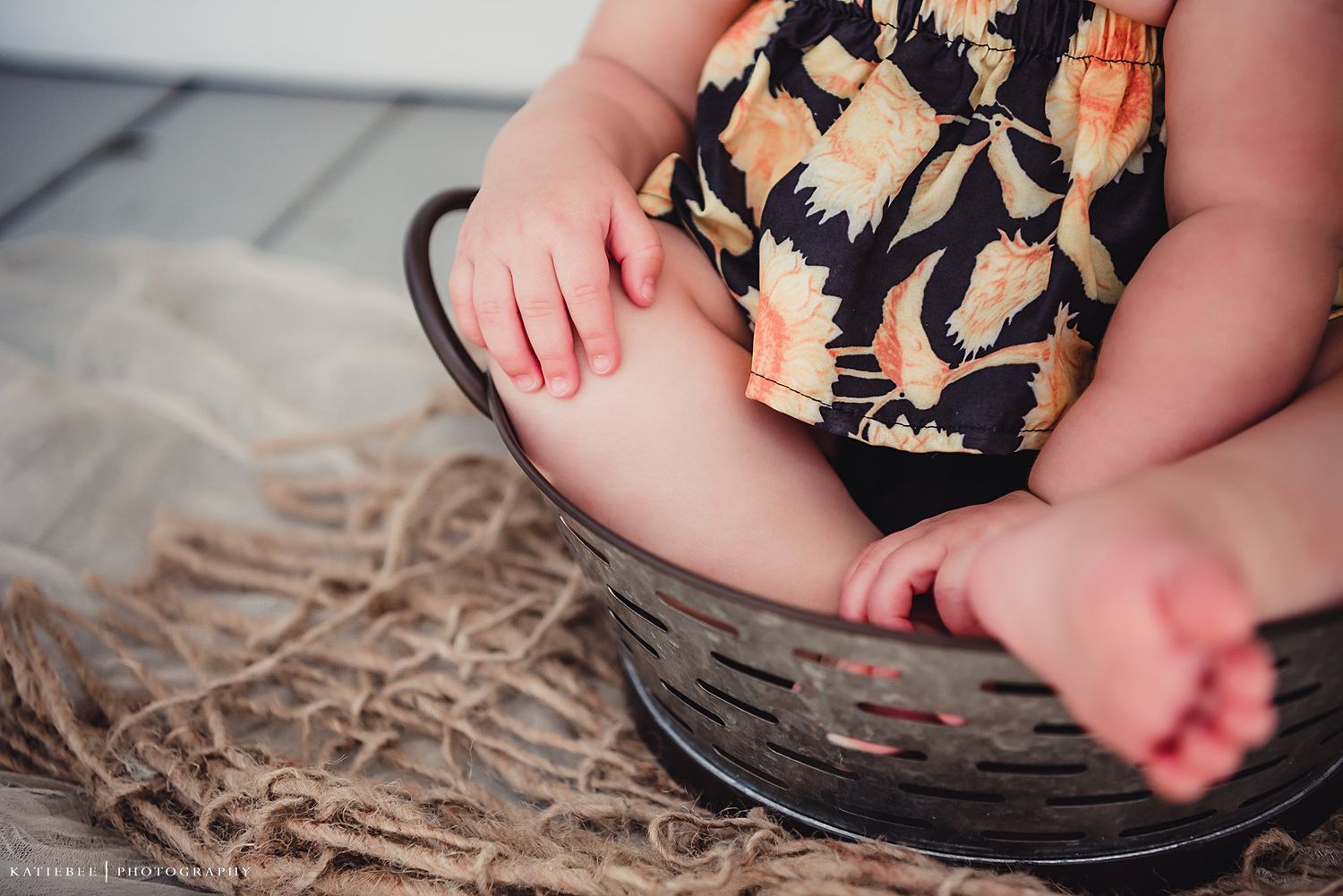 Baby legs in floral romper sitting in vintage brown basket with textured blanket.
