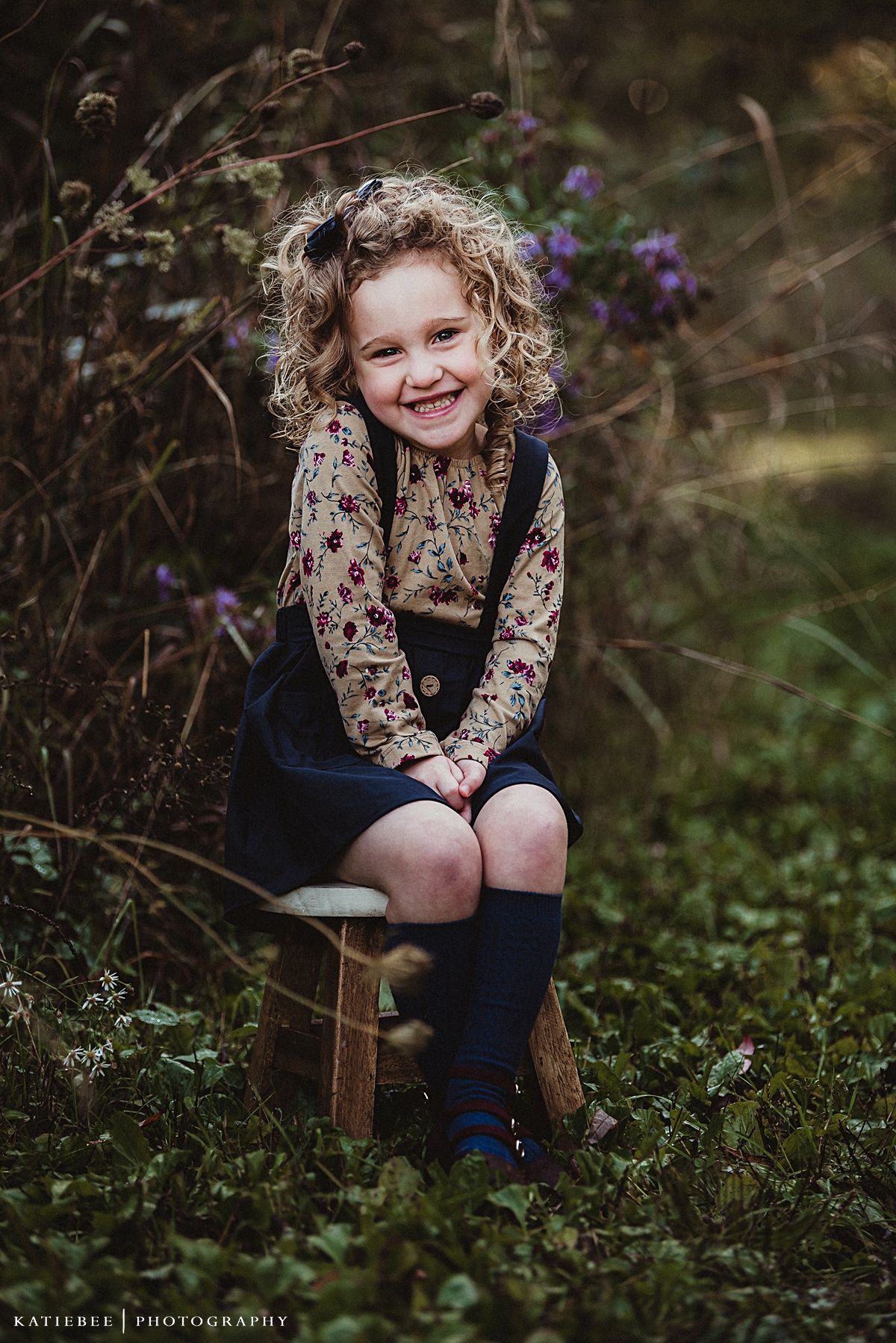 Child in floral top and navy suspender outfit sits on wooden stool among wild flowers.
