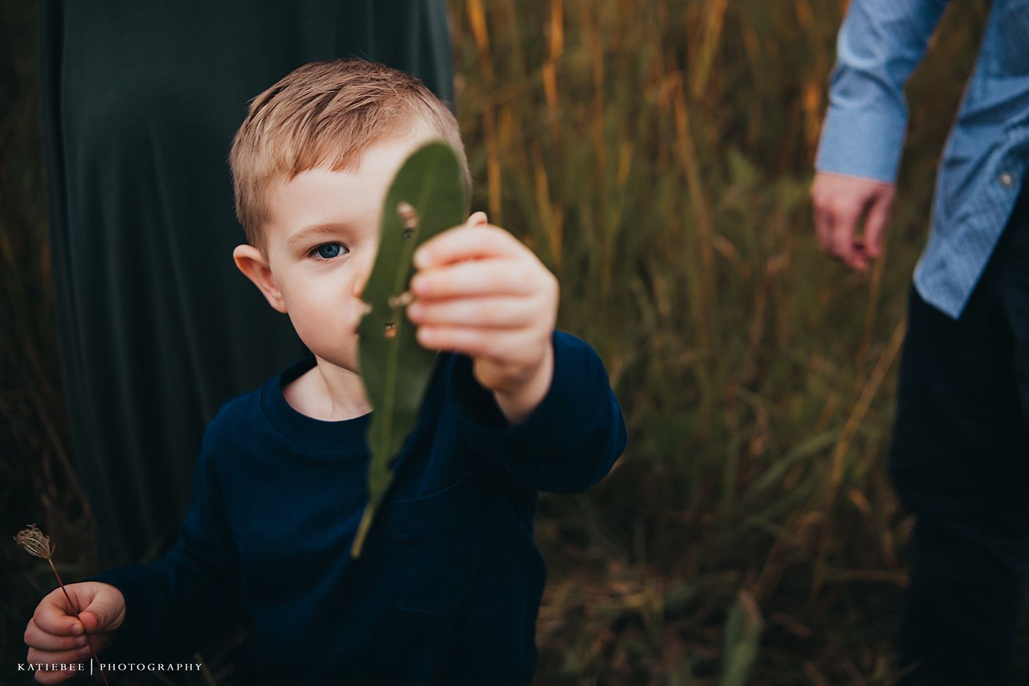 Child in navy sweater examining a leaf in tall autumn grass.