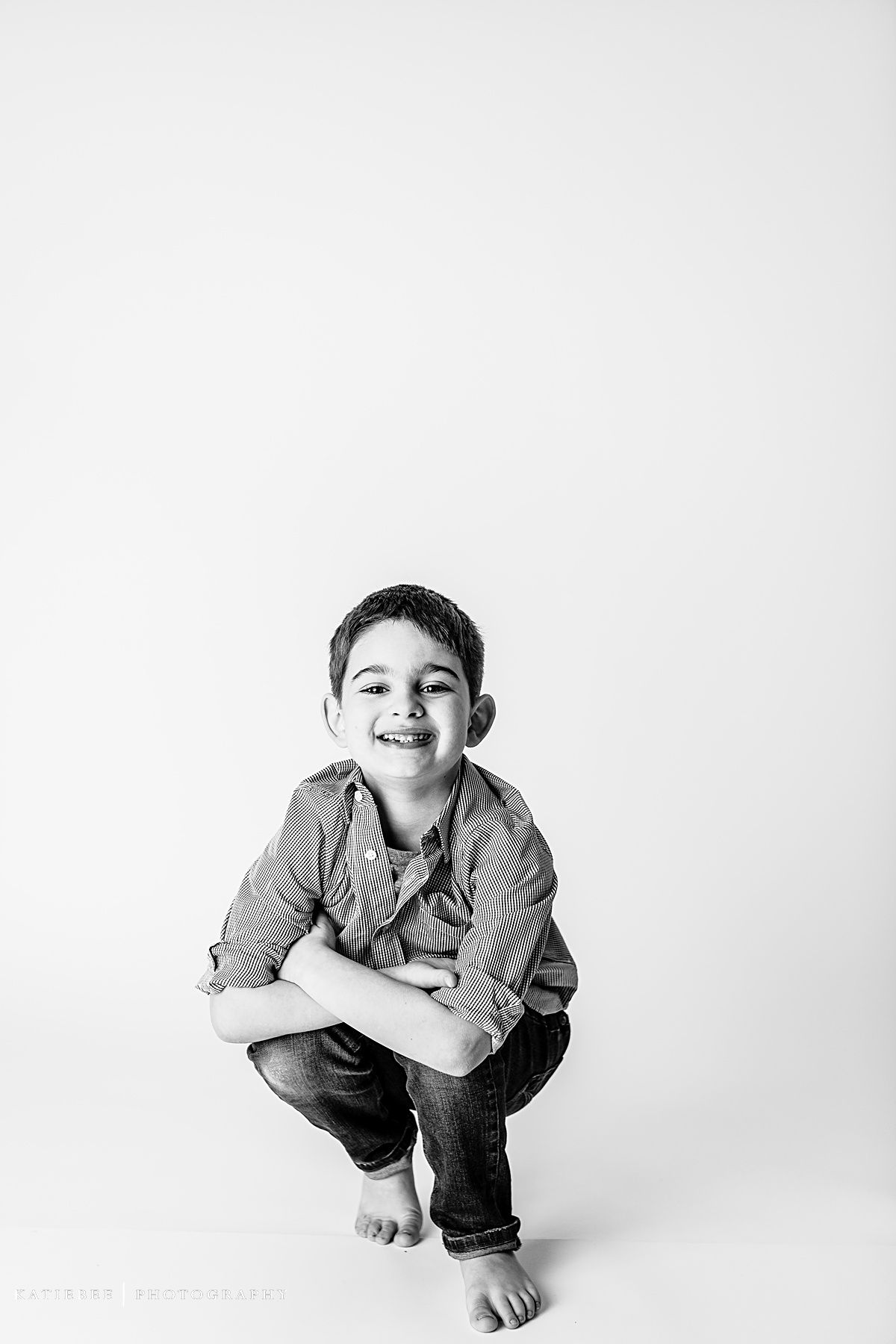Child crouching barefoot in casual attire against white backdrop in monochrome photo.