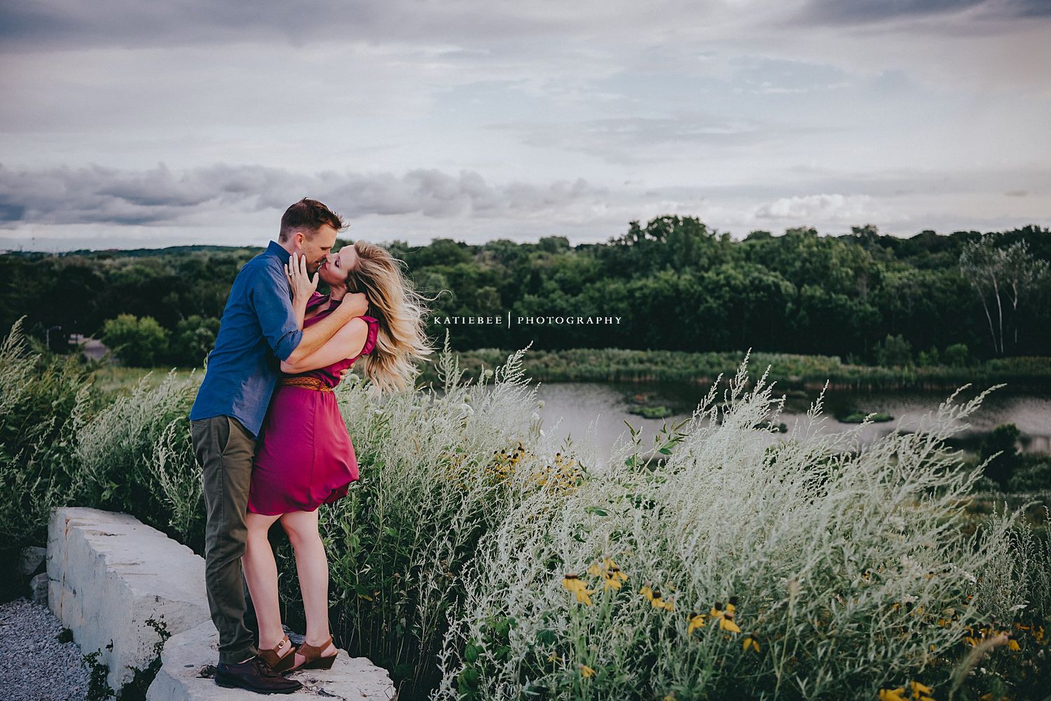A romantic couple embraces on a cliff overlooking a serene lake surrounded by lush greenery.