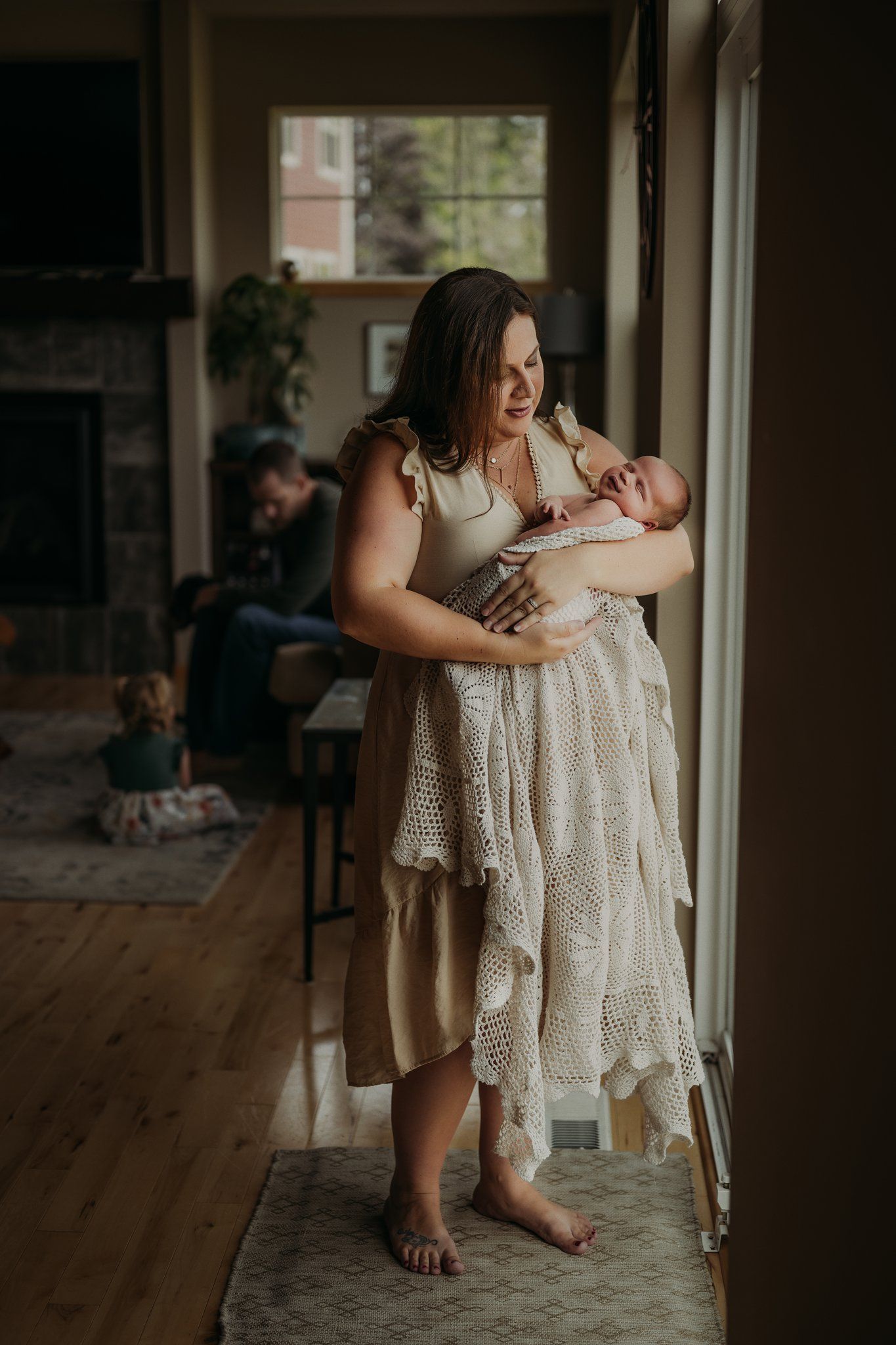 A tender moment captured by a window as a woman in a cream lace dress holds a newborn in natural light.