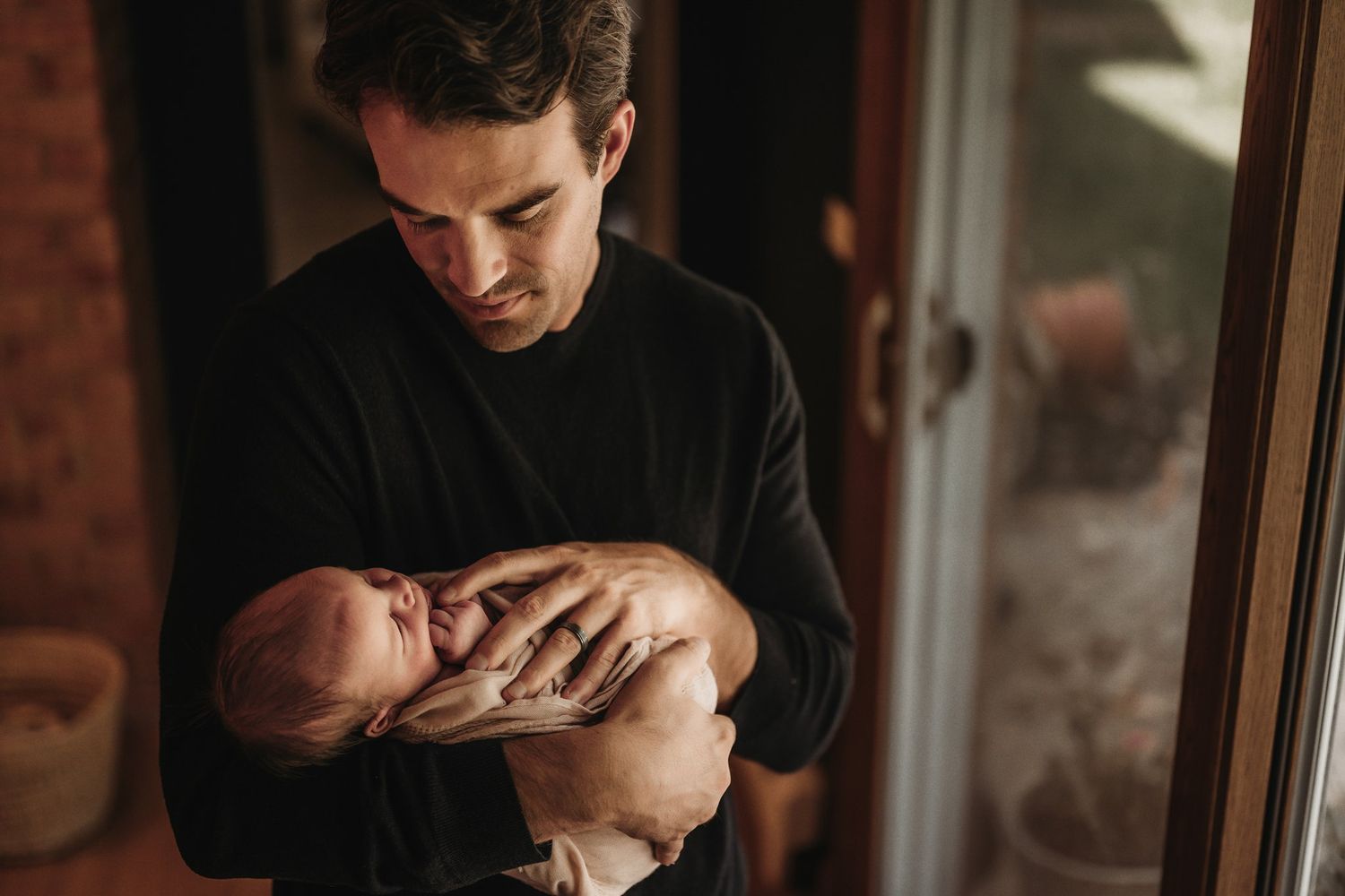 A tender moment as a person in black cradles a newborn baby near a window in warm lighting.