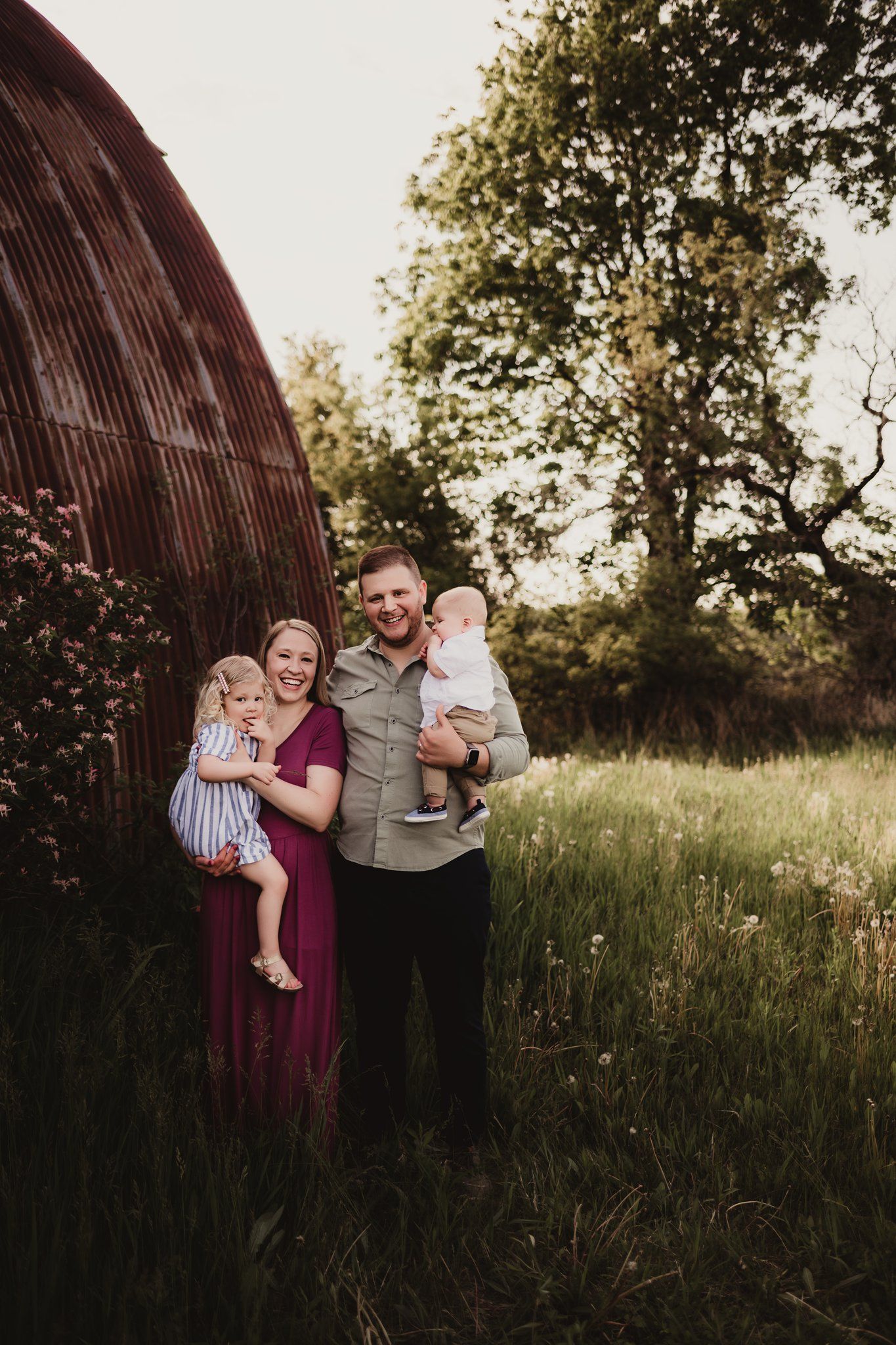 A family stands together near a rustic barn in a grassy field at sunset.