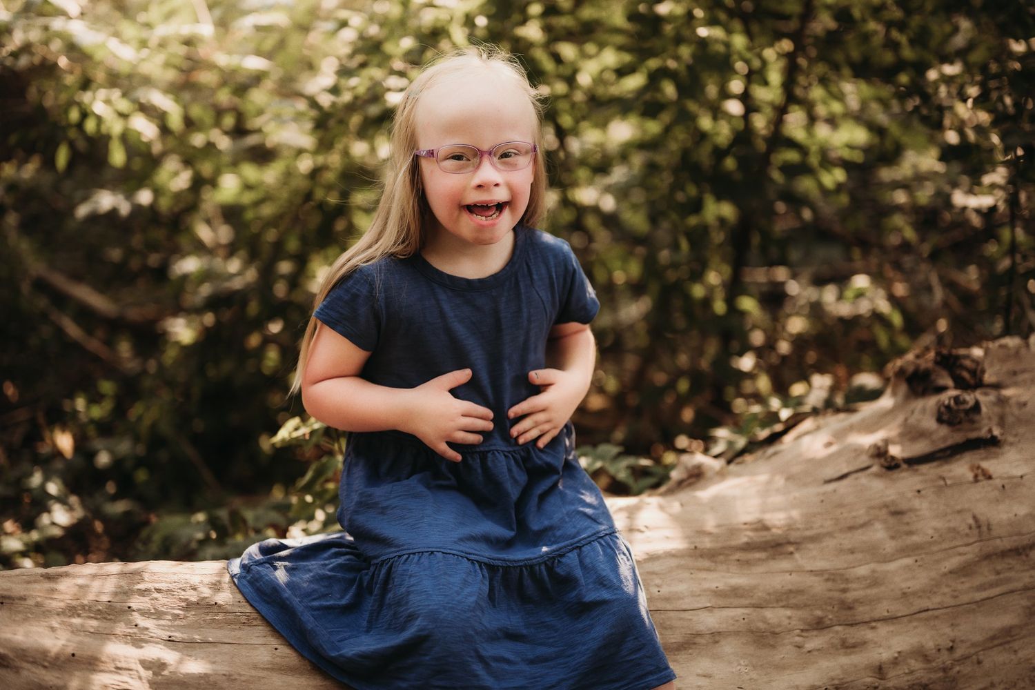 A joyful child wearing a navy blue dress sits on a fallen log in a sun-dappled forest setting.
