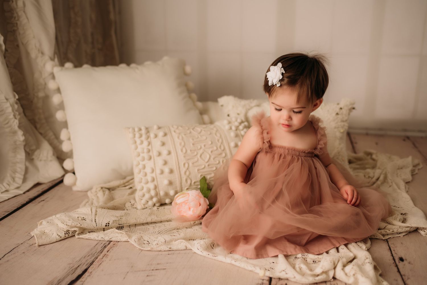 Baby sitting on textured cream blanket with decorative pillows in soft studio lighting.