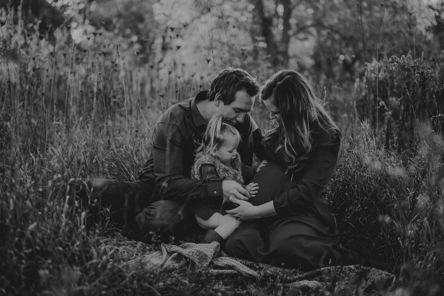 A couple shares an intimate moment with their child in a black and white outdoor photoshoot.