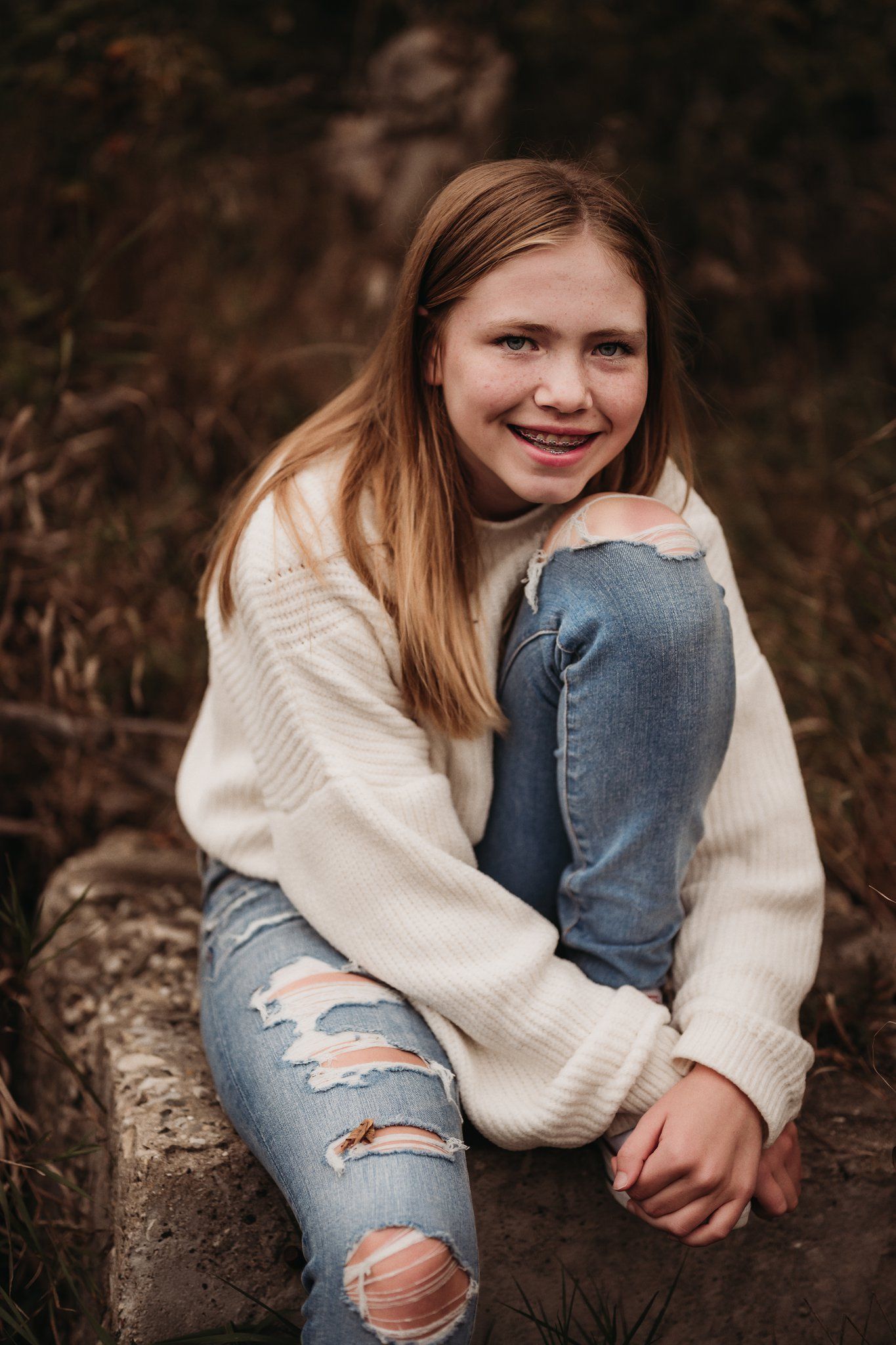 Person in cozy white sweater and ripped jeans sits casually on log outdoors.
