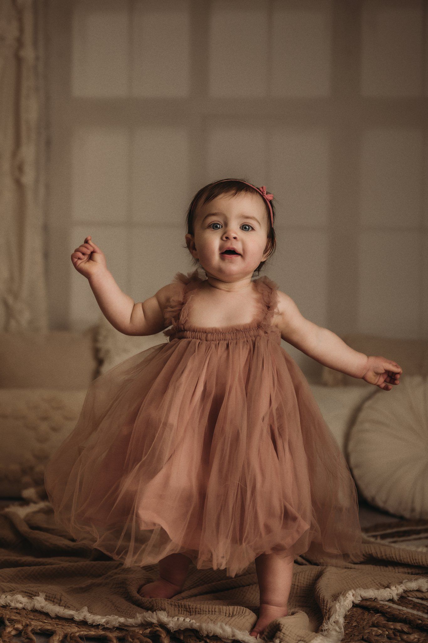 Small child in flowing mauve tulle dress standing against neutral interior backdrop.