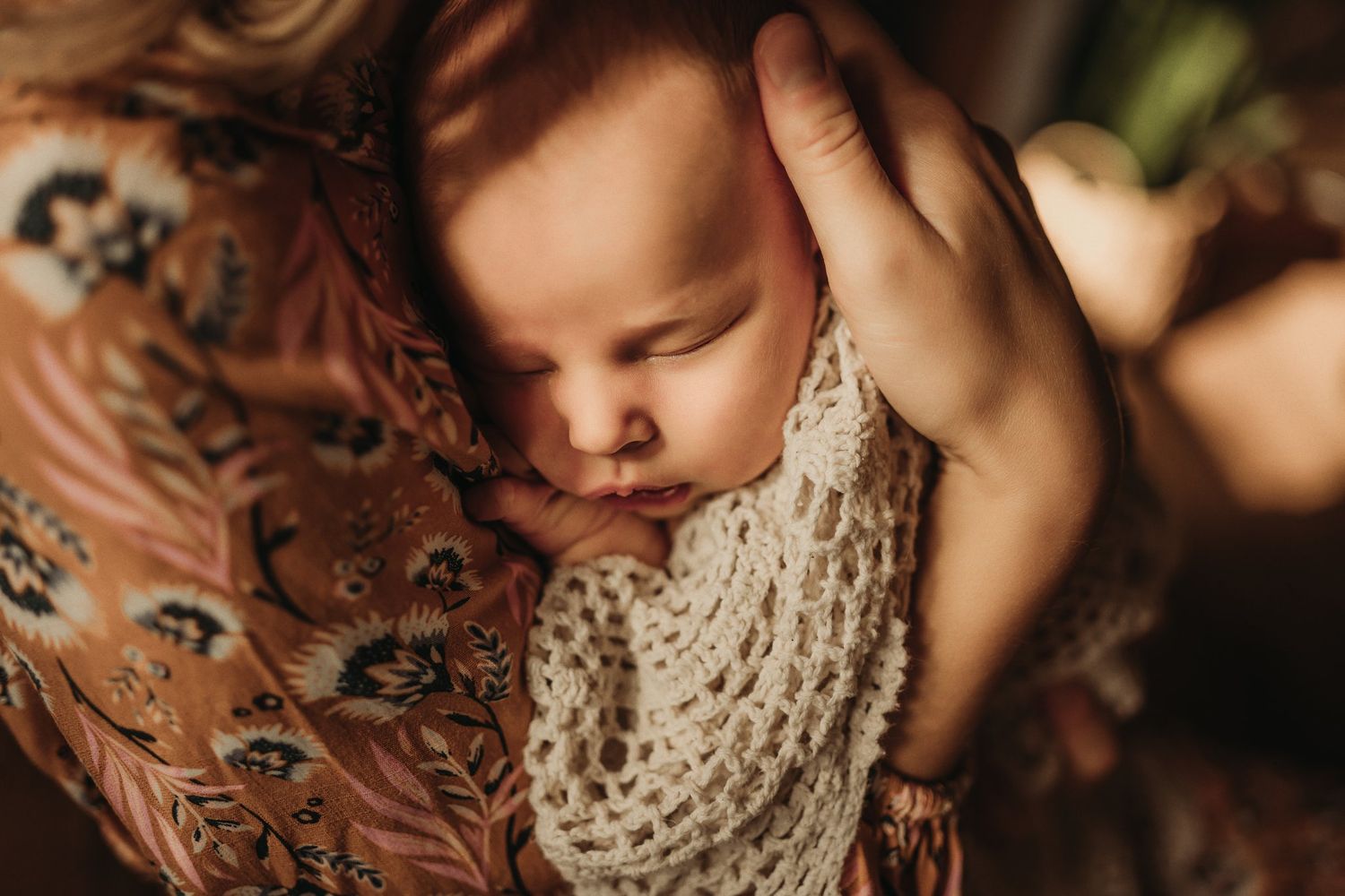 Close-up of a sleeping baby wrapped in a cream knit blanket against floral fabric.