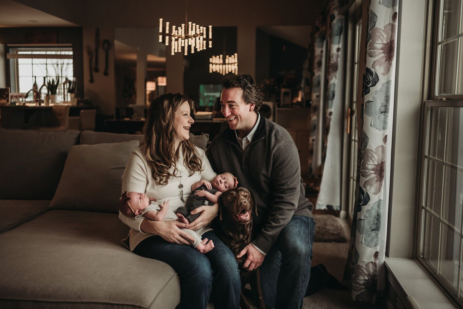 A cozy family moment on the sofa in a warmly lit living room with windows and neon sign in background.