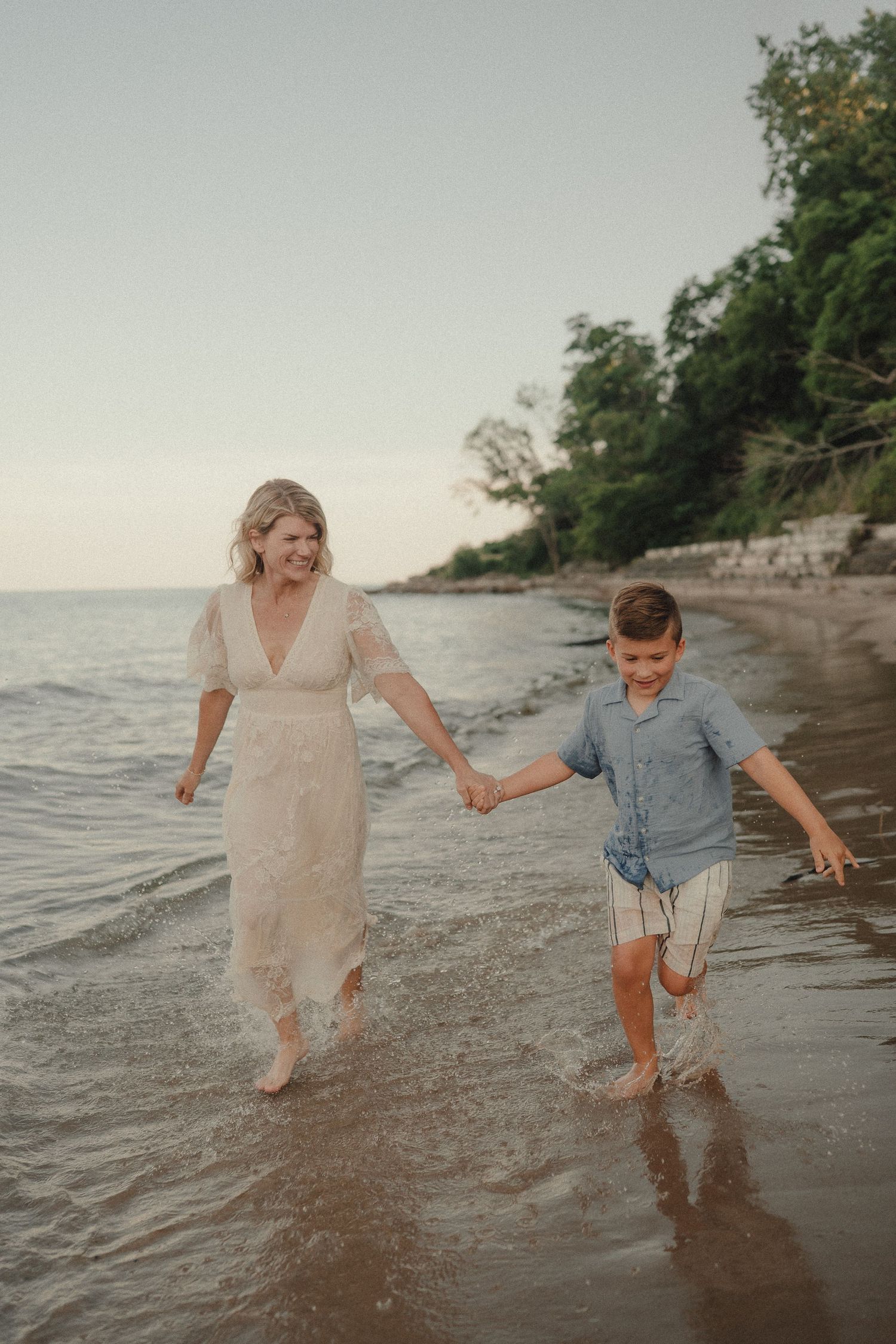 Smiling mother in white lace dress holding hands with her son splashing in ocean waves.