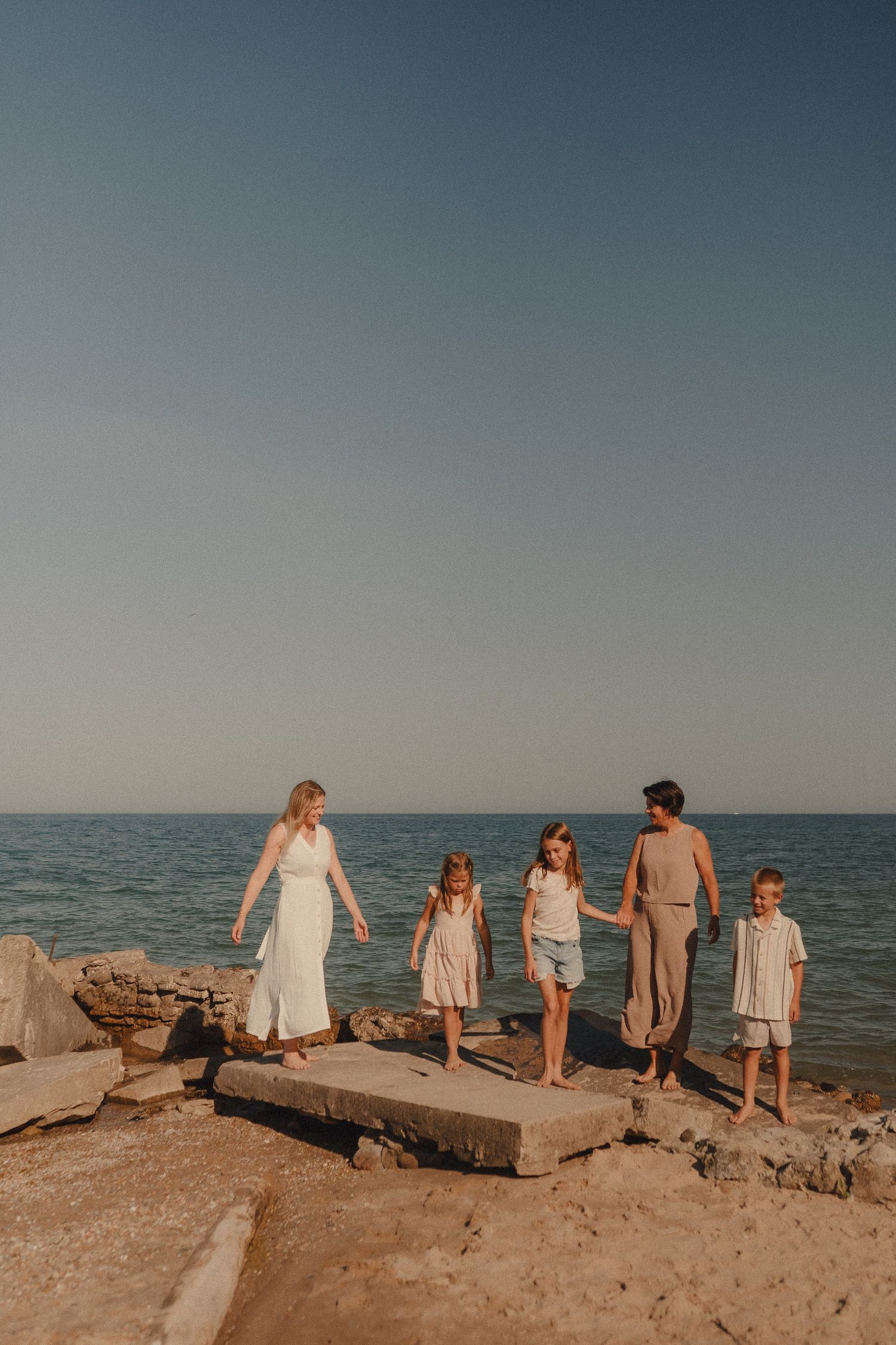 Two mothers and three children walking on rocky shoreline with calm blue ocean backdrop.