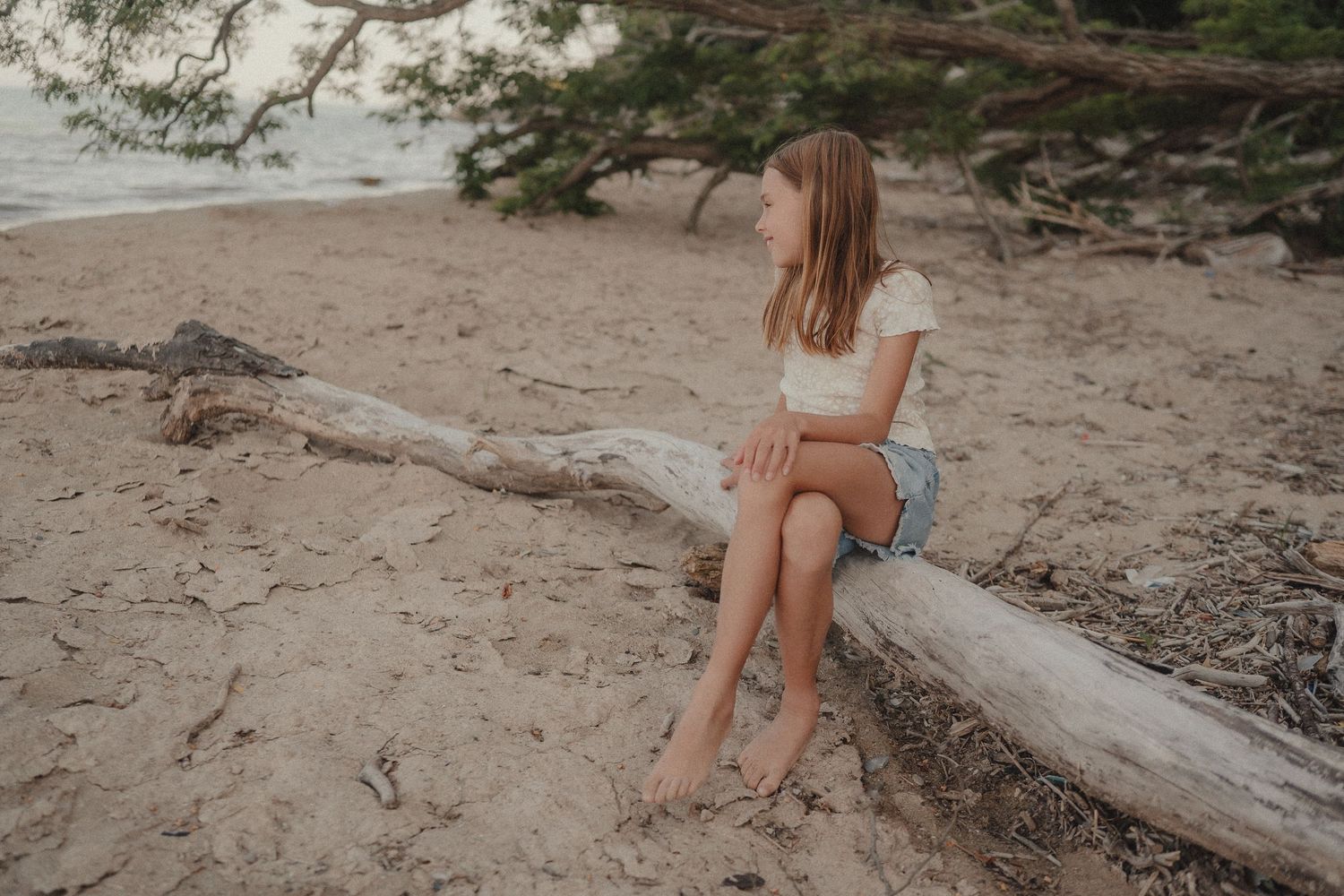 A barefoot girl sits on a driftwood log on a sandy beach, gazing toward the ocean surrounded by pine trees.