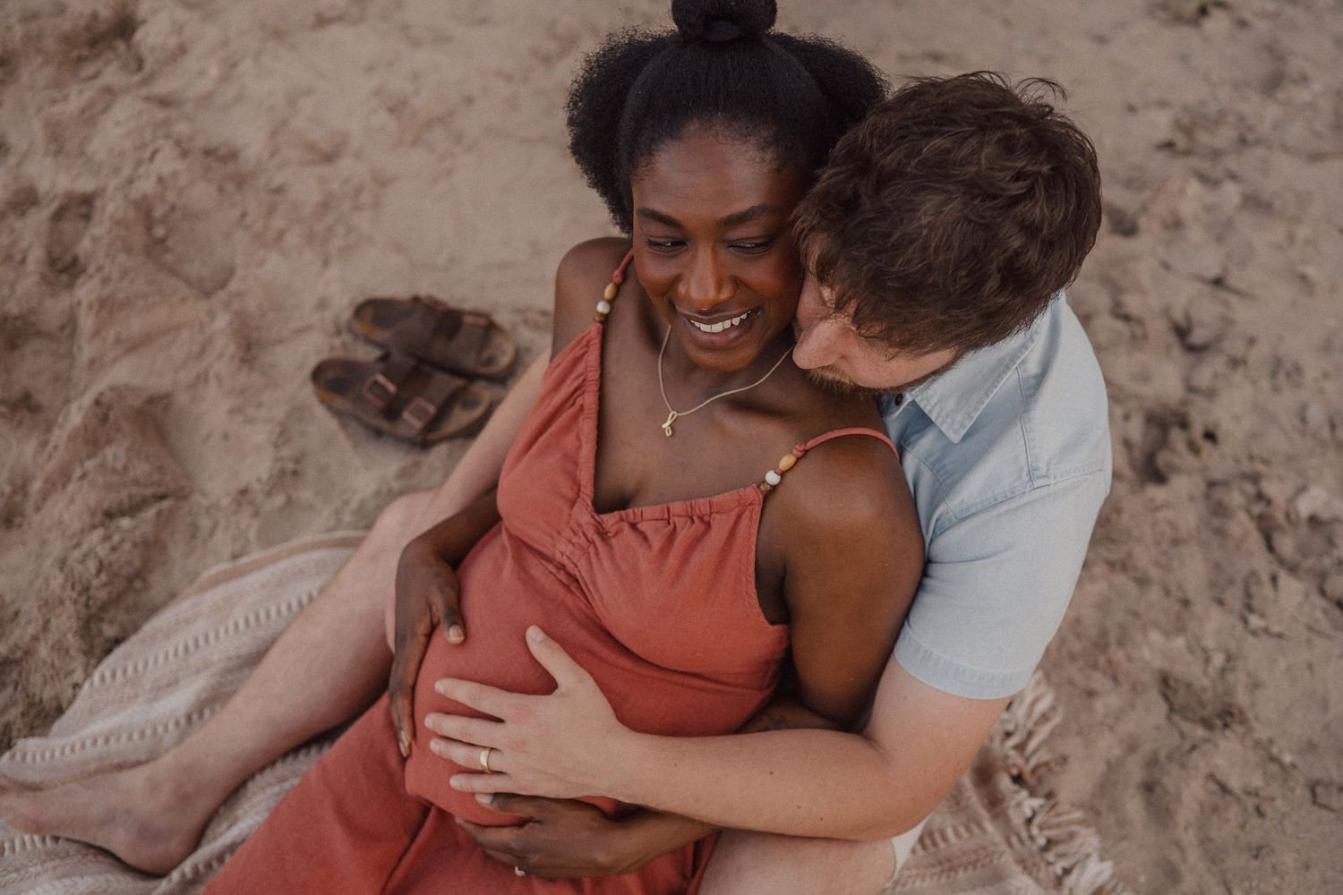 Couple sitting on beach blanket in sand, woman in coral dress smiling as man embraces her from behind.