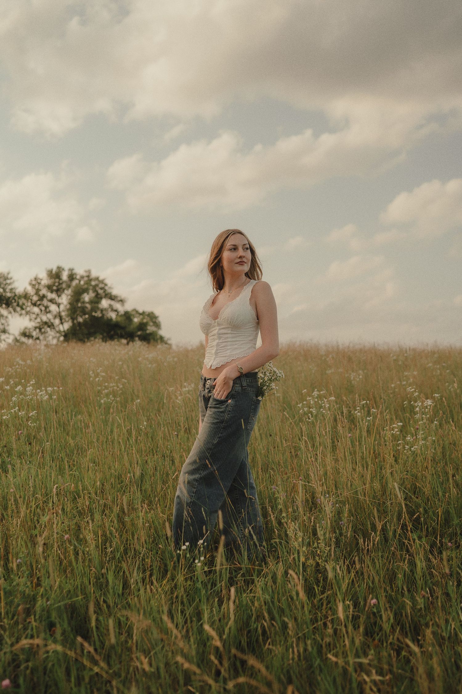 Young woman in white tank and jeans stands in tall wildflower meadow under cloudy sky.