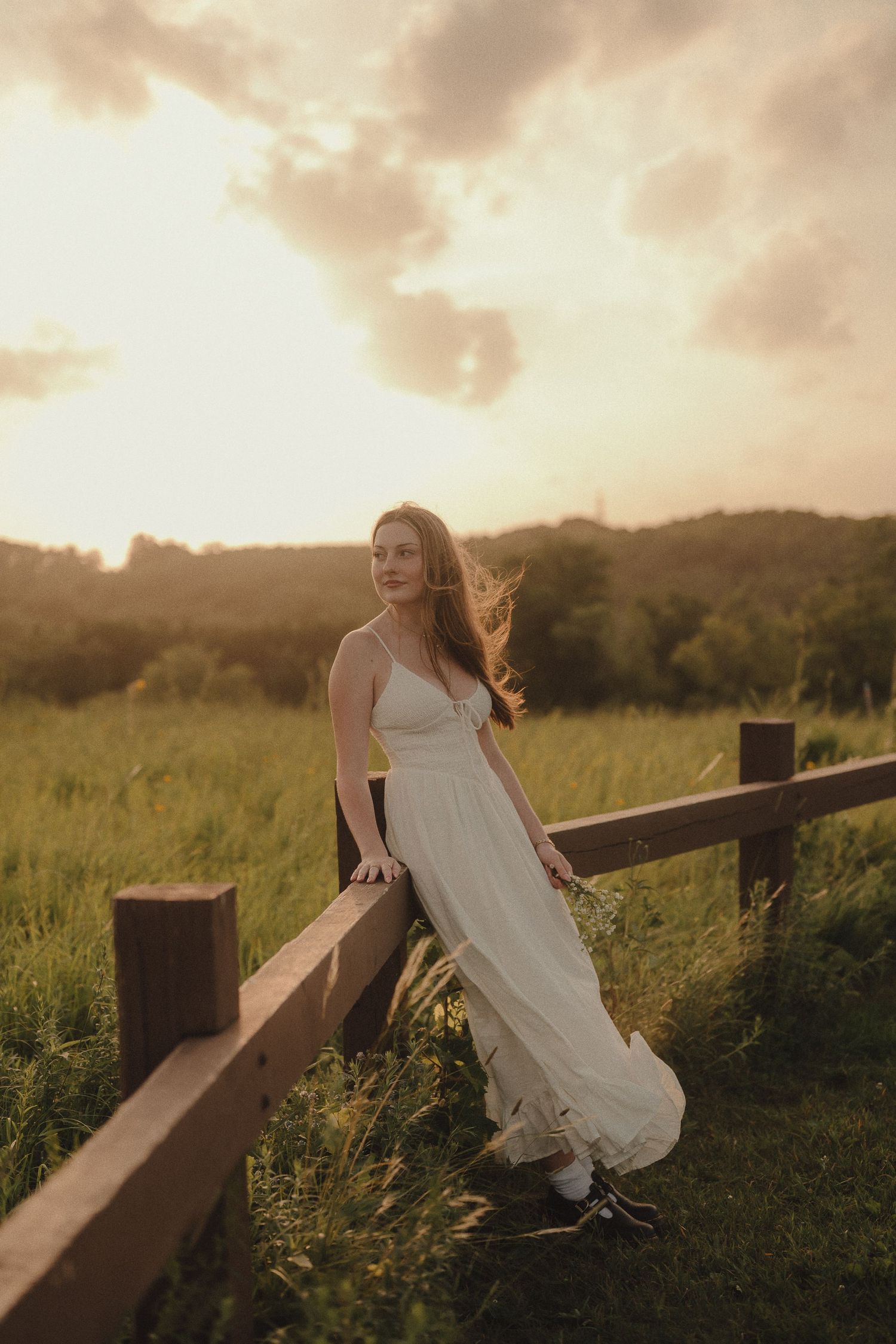 Young woman in flowing white dress stands by wooden fence in green meadow at sunset.